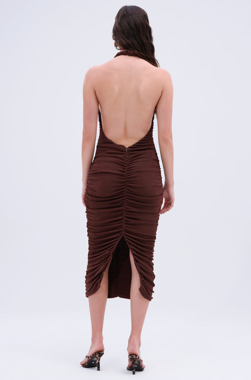 A person, with wavy hair cascading freely, stands facing away from the camera to showcase the BODY ODDY MIDI DRESS IN BROWN. This ruched midi dress features a halter neckline and a bottom slit, paired with high-heeled sandals against a simple, light-colored backdrop.