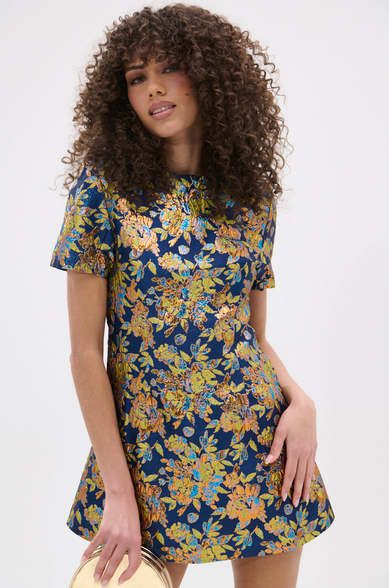 Woman with curly hair wearing the BLUE IS THE NEW BLACK BROCADE MINI DRESS, featuring a gold and yellow floral pattern, holding a small round gold purse against a plain light background.