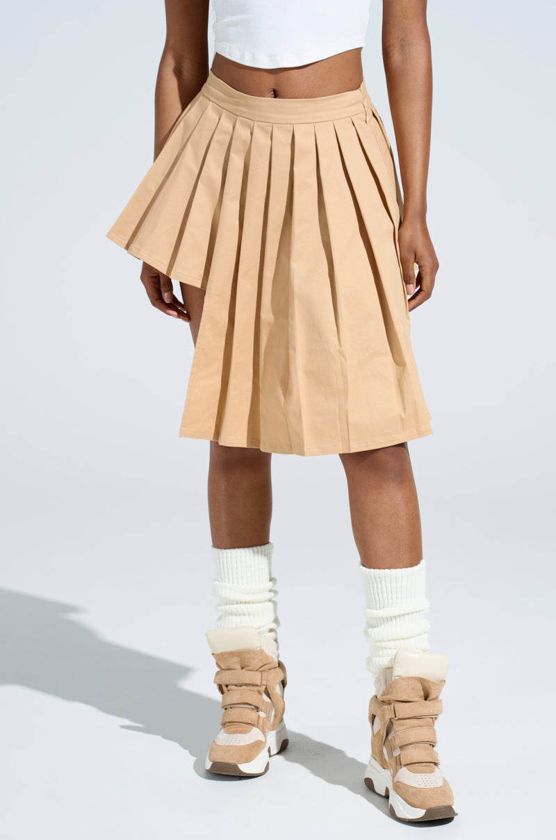 A person confidently poses in a SHARI BABY PLEATED MINI SKIRT with an asymmetrical hem, paired with a white cropped top and high-top beige sneakers. White socks are scrunched down over the sneakers, all set against a plain background.