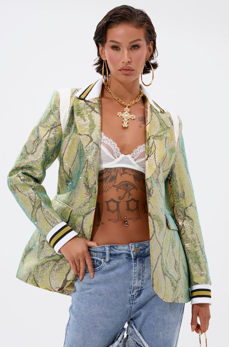 A confident woman models the DON'T TRY ME EMBELLISHED BROCADE BLAZER in metallic green over a white lace bralette, showcasing torso tattoos, large hoop earrings, a gold cross necklace, blue jeans, and wet-styled hair.