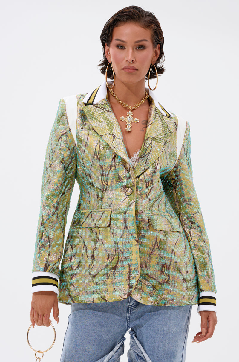 A confident woman wears the DON'T TRY ME EMBELLISHED BROCADE BLAZER in metallic green with white and black striped cuffs and shoulder accents, paired with layered necklaces, hoop earrings, ripped blue jeans, and a gold ring accessory.