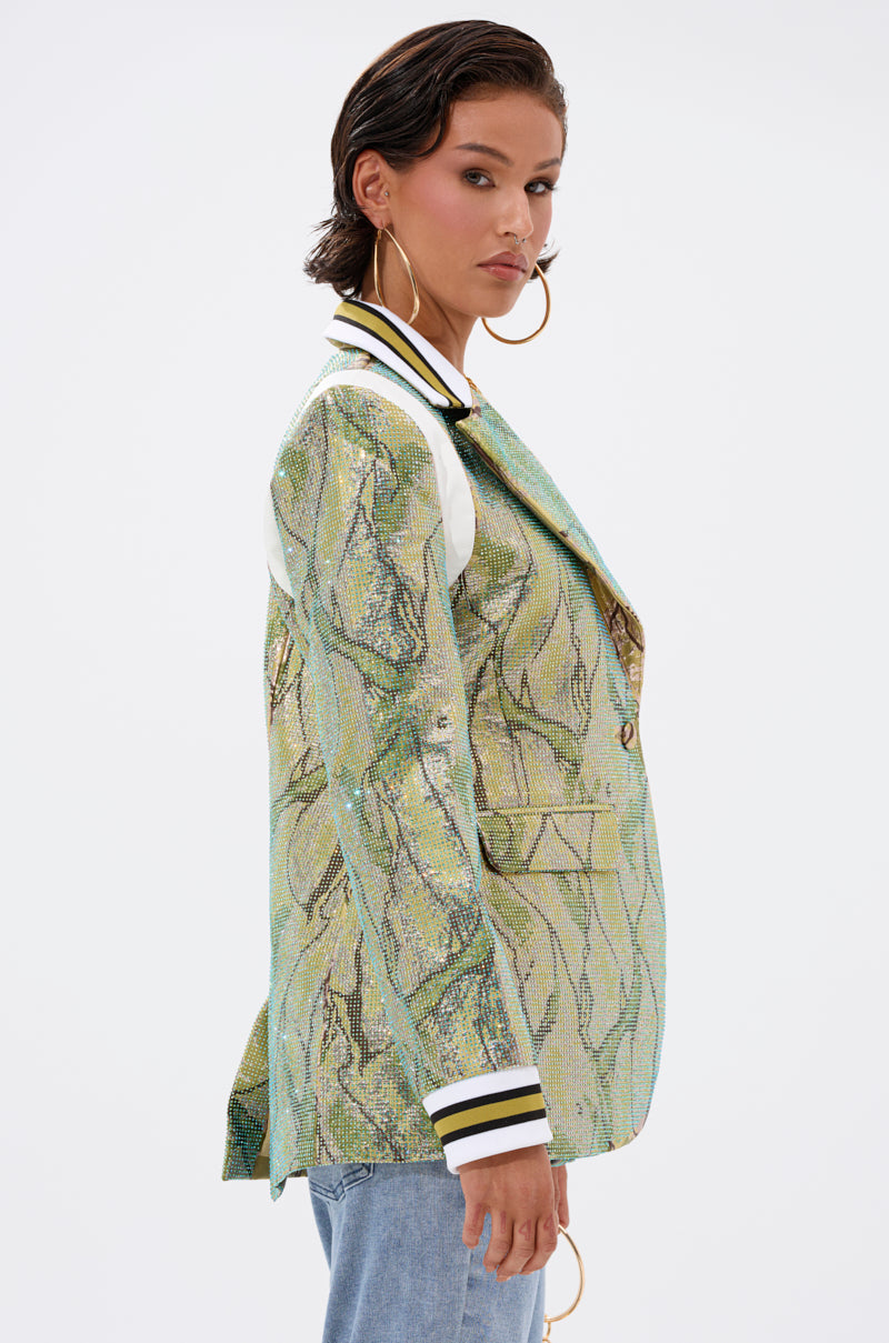 A woman with short dark hair models the DON'T TRY ME EMBELLISHED BROCADE BLAZER, featuring a metallic green finish and black-and-white striped collar and cuffs, paired with large hoop earrings and light blue jeans against a plain white background.