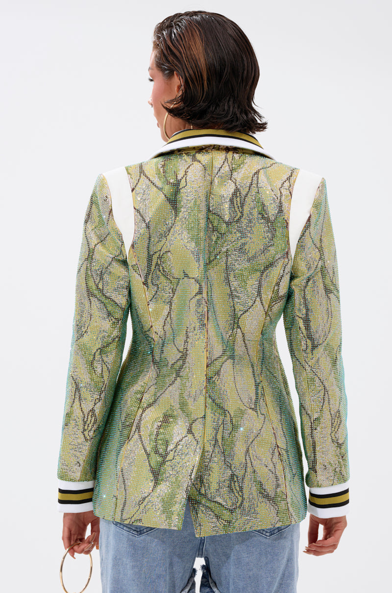 A person with short, slicked-back hair is shown from behind wearing the DON'T TRY ME EMBELLISHED BROCADE BLAZER, a green statement piece with white and black striped accents, and holding a circular gold accessory in their left hand.