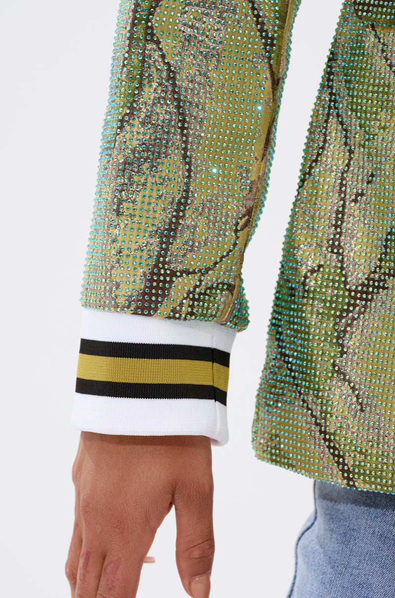 A close-up of a person's arm in the DON'T TRY ME EMBELLISHED BROCADE BLAZER, featuring green jewel-studded forest patterns, a white cuff with black and yellow stripes, paired with light blue jeans.