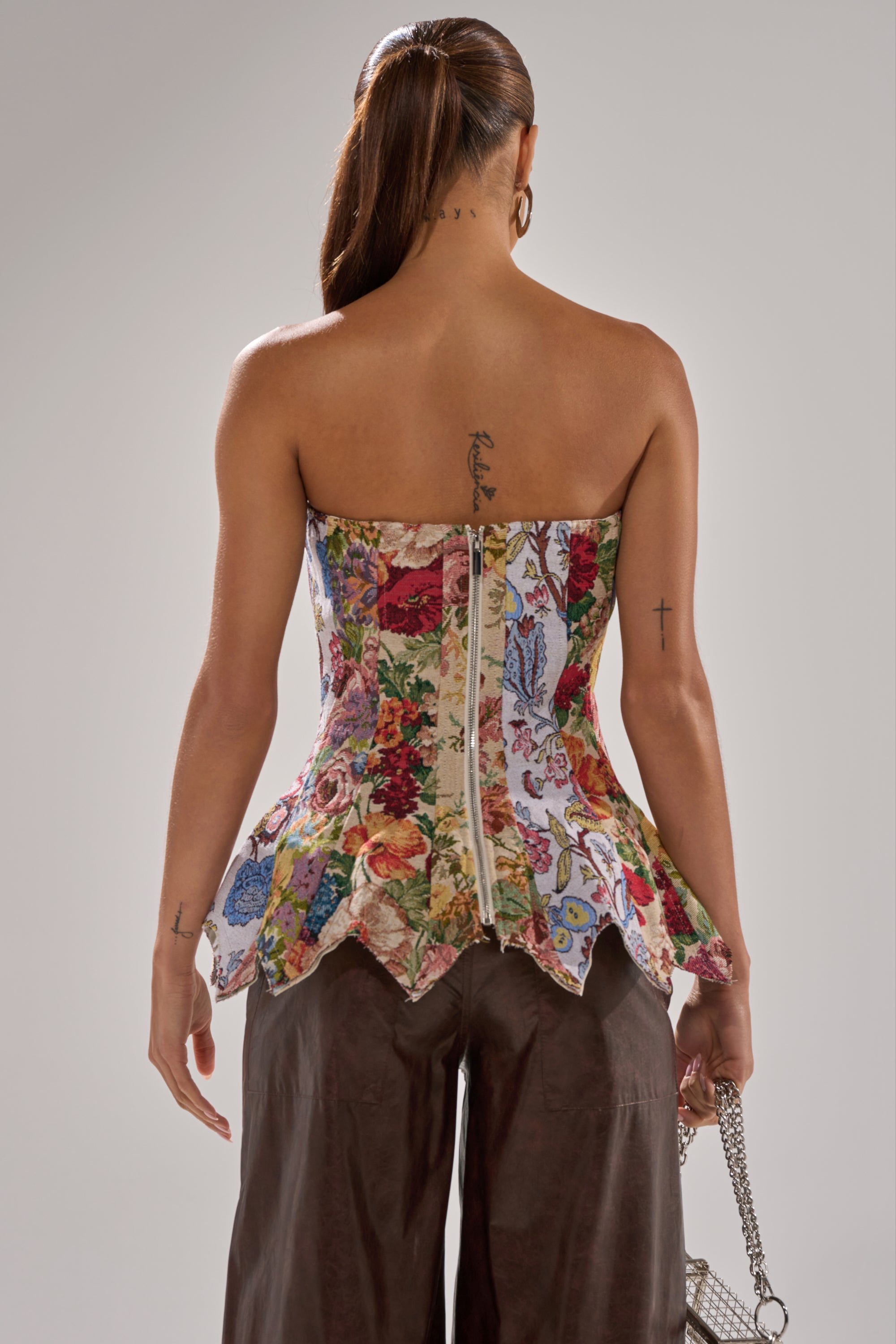 A woman faces away from the camera, wearing the SUNDAY PATCHWORK CORSET peplum top and brown pants. She holds a small silver handbag, with visible tattoos on her upper back and arms.