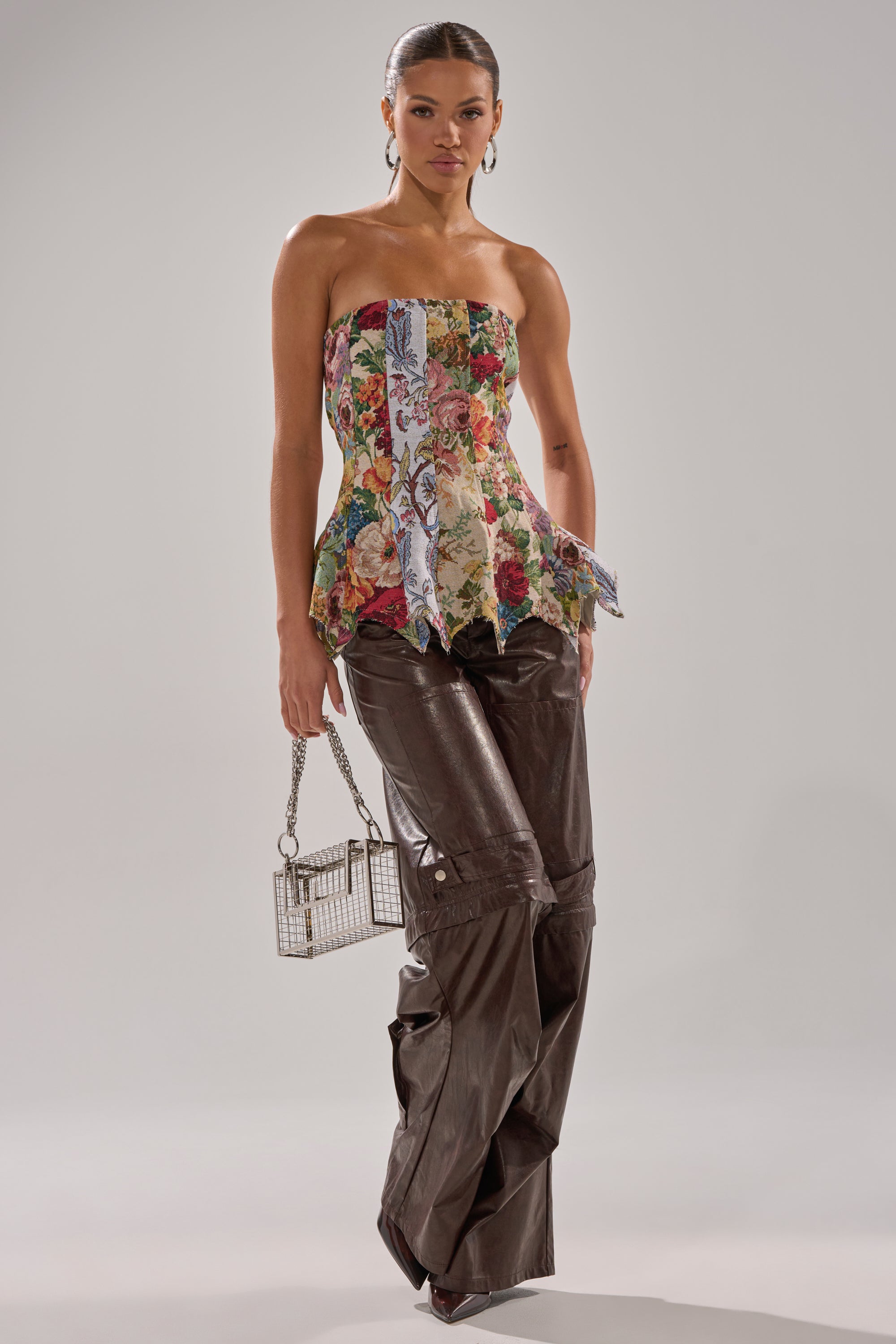 A woman models the SUNDAY PATCHWORK CORSET with a floral handkerchief hem, styled with brown wide-leg leather pants, a metallic mini handbag, hoop earrings, and her hair pulled back.