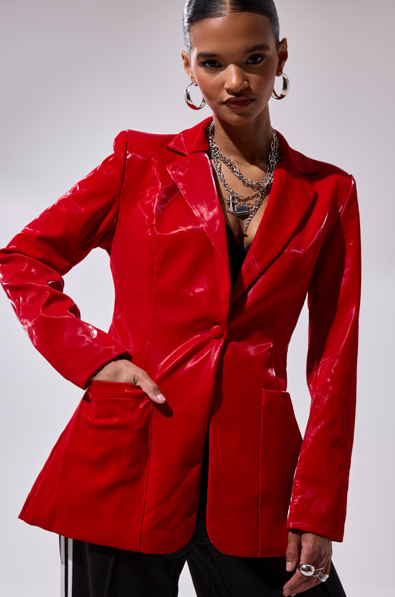 A woman poses confidently against a plain background wearing the TOO SLICK LIQUID VELVET BLAZER, featuring a shiny faux patent leather finish and large pockets, styled with layered silver necklaces, hoop earrings, and black pants.