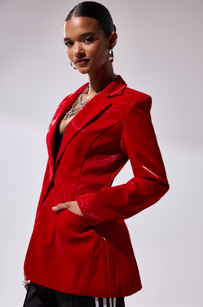 A woman with slicked-back hair poses confidently in the TOO SLICK LIQUID VELVET BLAZER, a glossy red faux patent leather piece, styled with statement earrings and layered necklaces against a plain light background.