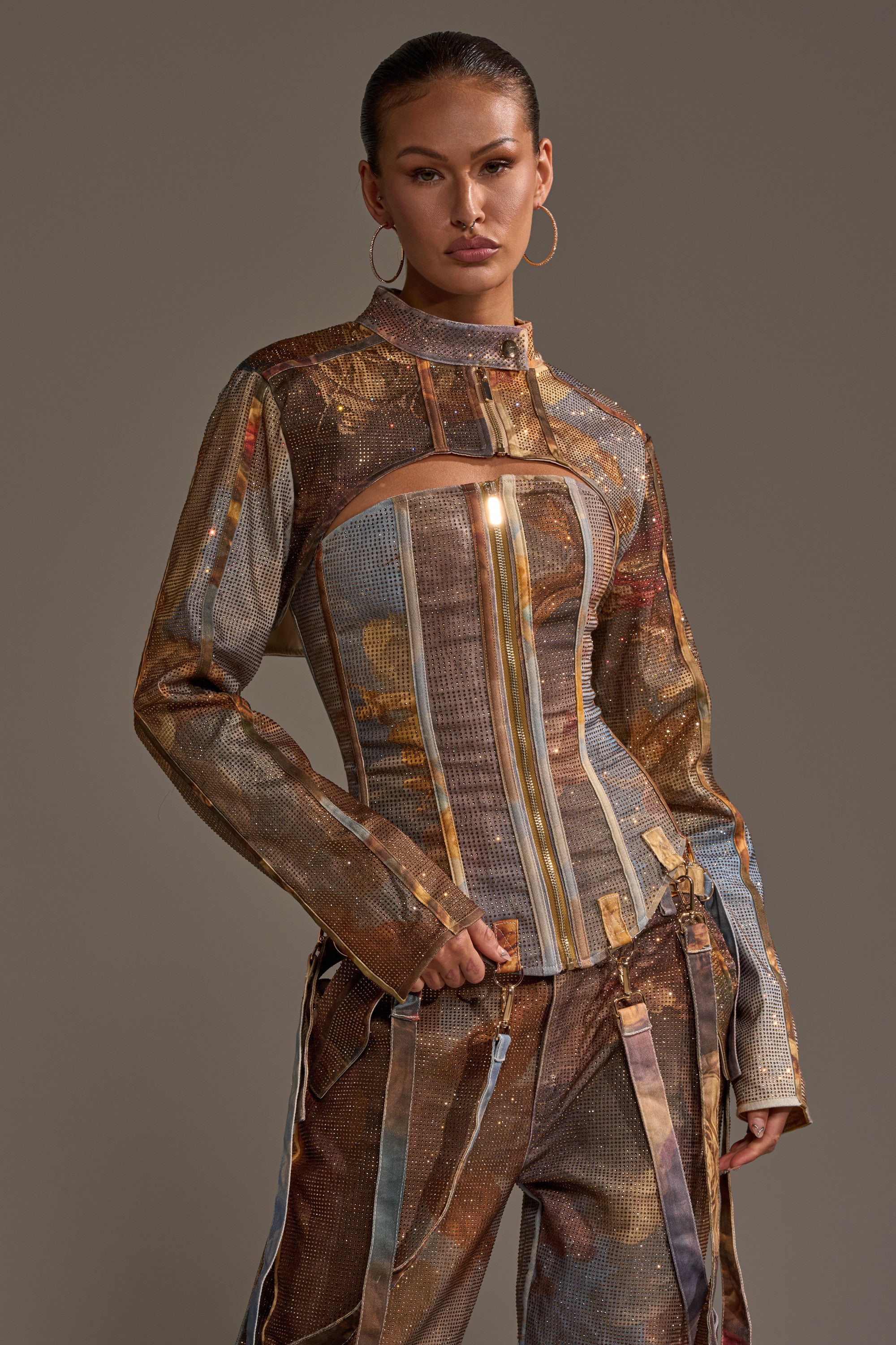 A woman models the IN THE FUTURE RHINESTONE BOLERO with a metallic, multicolored outfit of a structured chest-cutout top and striped pants. She accessorizes with hoop earrings and slicked-back hair against a neutral background.