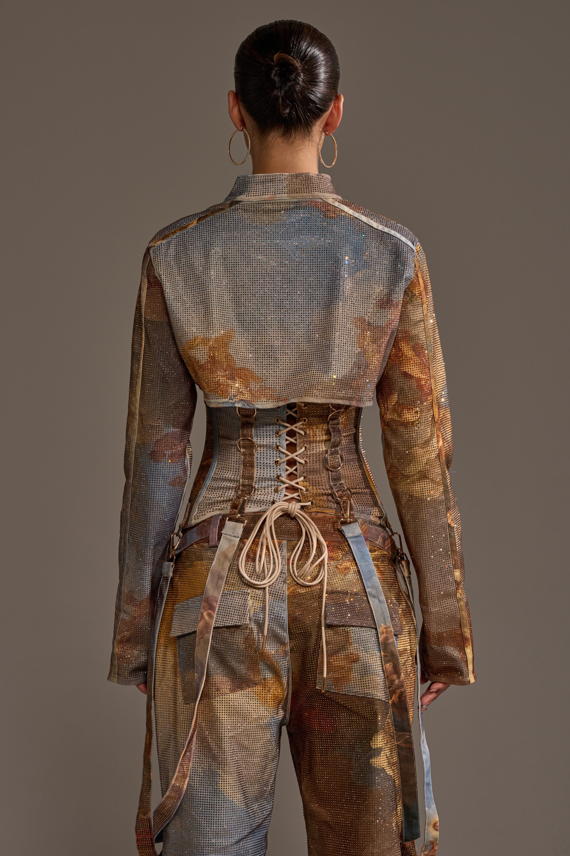 A person with dark hair in a bun faces away, wearing the shimmery IN THE FUTURE RHINESTONE BOLERO with graphic print, lace-up corset back detail, and large hoop earrings against a plain background.