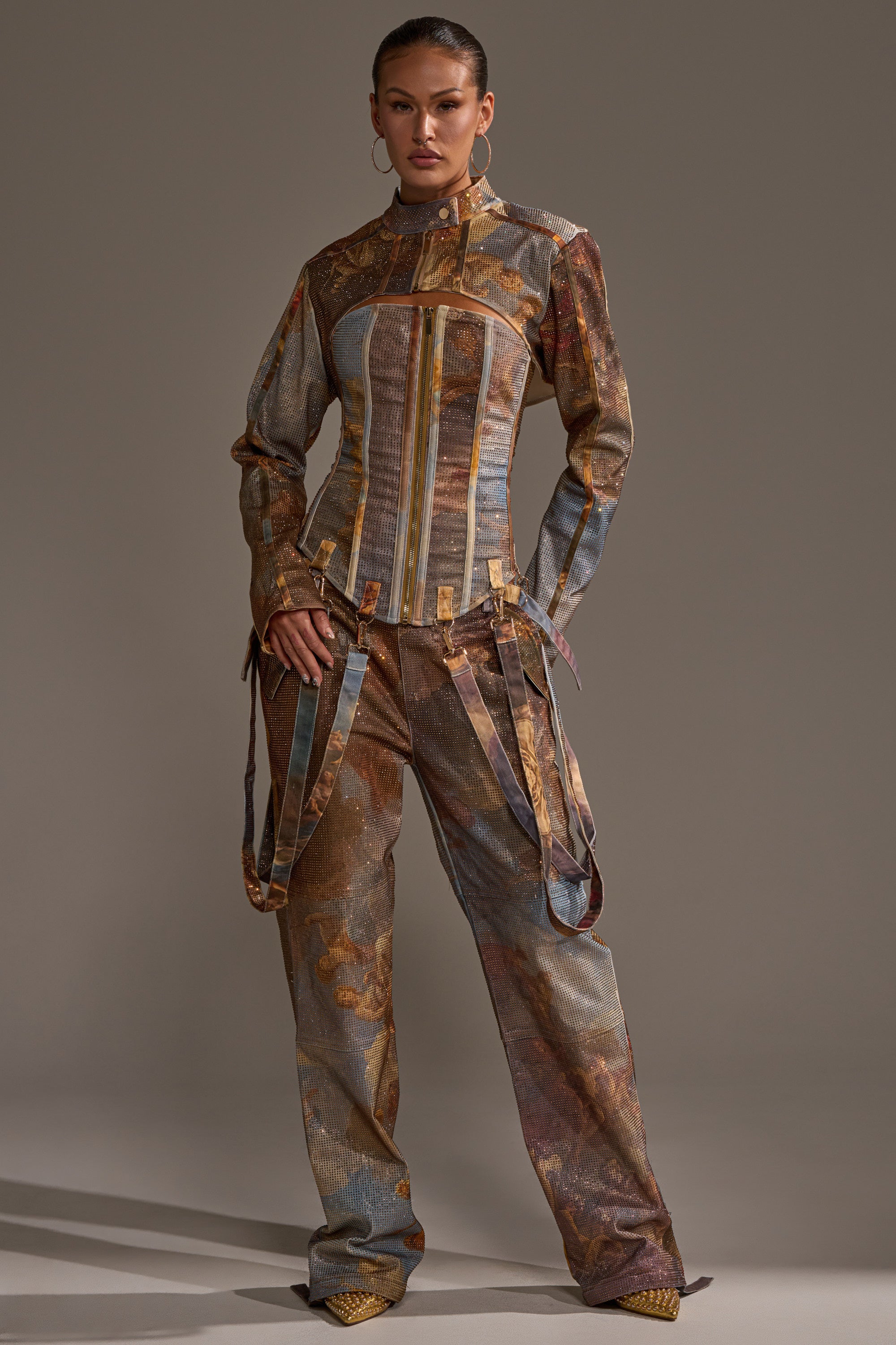 A woman stands confidently in the IN THE FUTURE RHINESTONE BOLERO, a graphic print jacket with earth-toned patterns, gold accents, and decorative straps, paired with matching wide-leg pants against a plain gray background.
