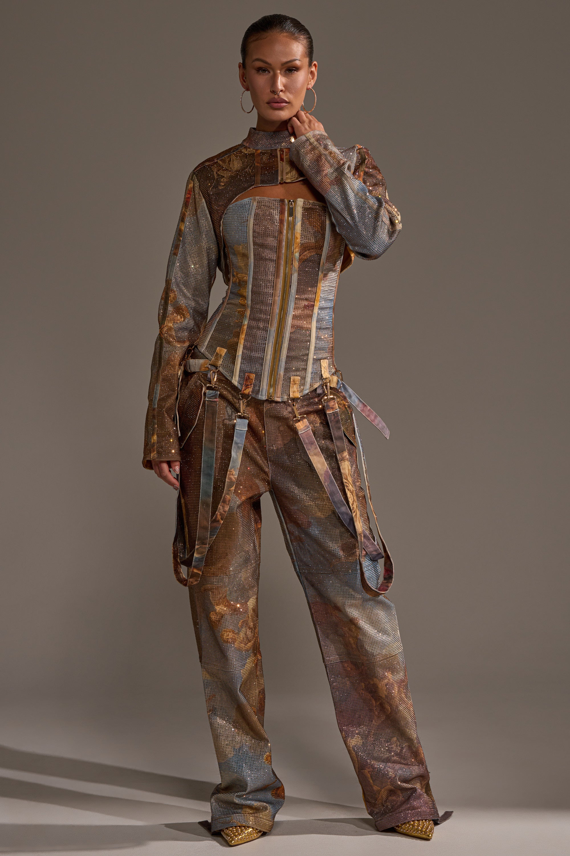 A woman stands confidently in a structured, earth-toned outfit with belted pants and a corset top, layering the IN THE FUTURE RHINESTONE BOLERO over her look; her gold shoes shine as she poses against a plain backdrop.