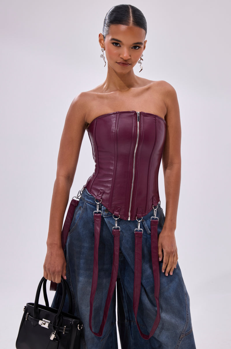 A woman wears the FAN FAVORITE FAUX LEATHER CORSET in burgundy with a front zipper and harness straps, paired with wide-leg blue jeans, a black handbag, and her hair slicked back.