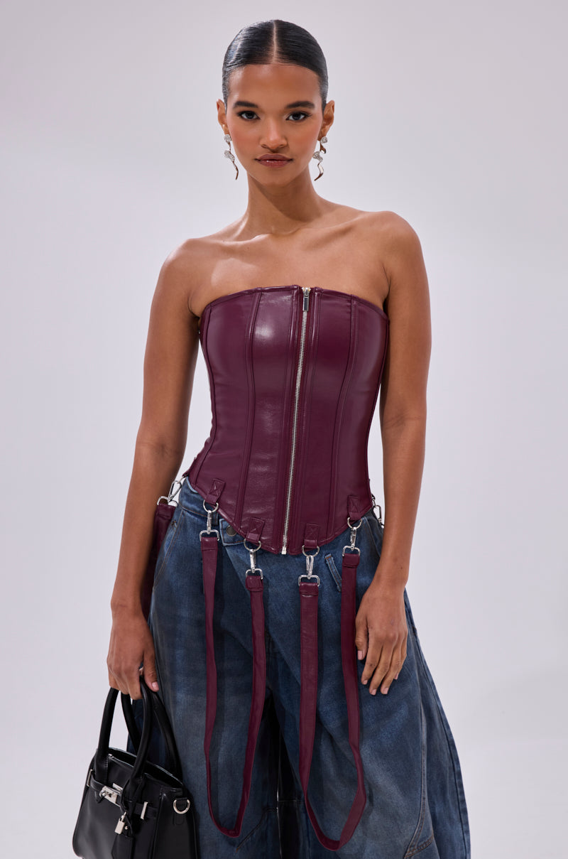 A woman models the FAN FAVORITE FAUX LEATHER CORSET with harness-style straps and zip closure, paired with wide-leg blue jeans and a black handbag, posing against a light gray background.