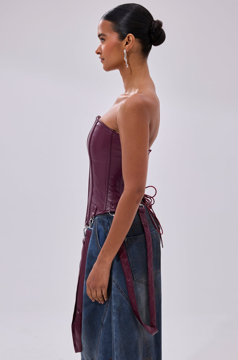 A woman stands in profile against a plain background, wearing the FAN FAVORITE FAUX LEATHER CORSET with harness-style straps and a long, dark denim skirt. Her hair is styled in a neat bun and she wears dangling earrings.
