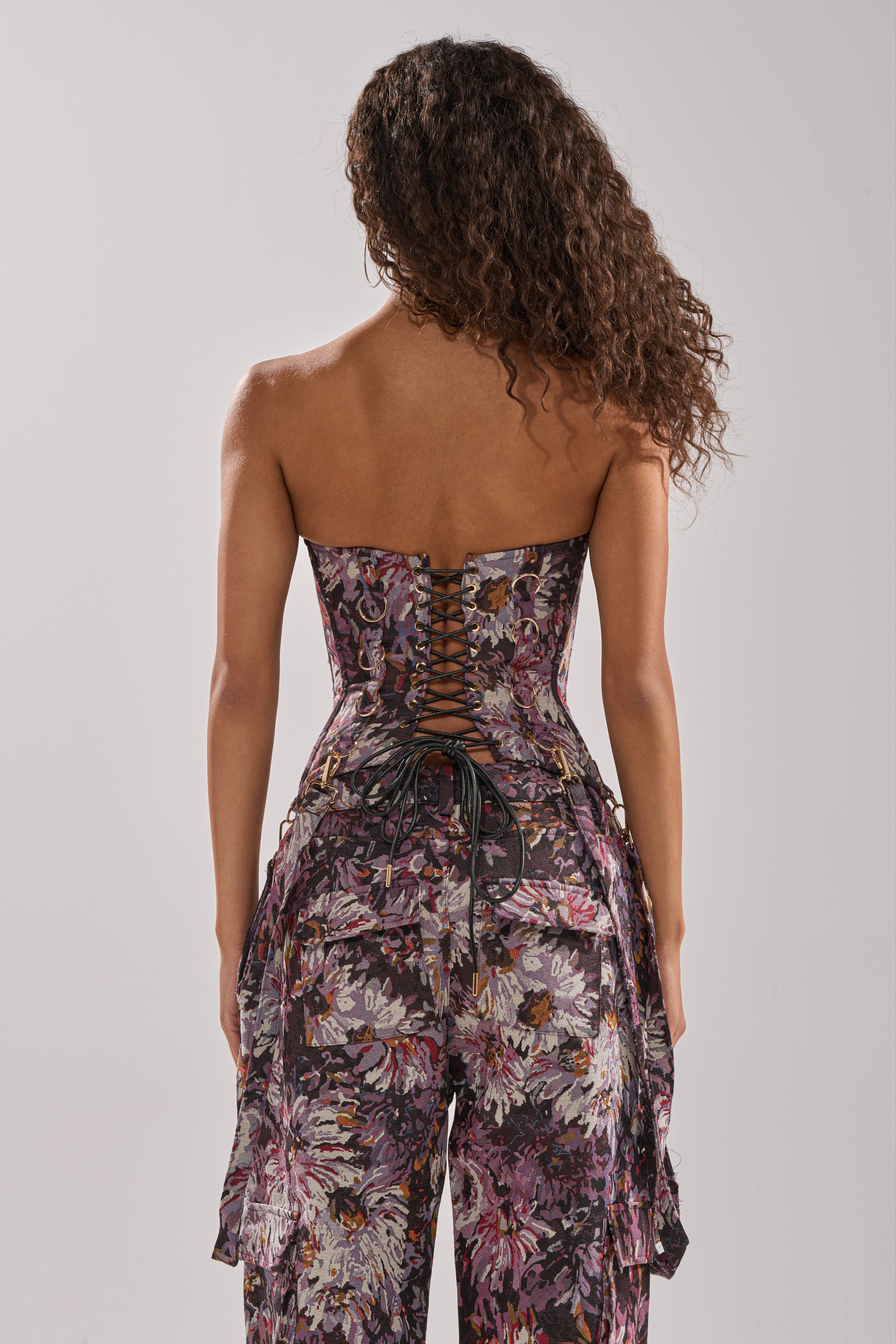 A woman with curly hair faces away, wearing the ABOVE THE MIDDLE FLORAL CORSET with a strapless design, corset-style lace-up back, and purple, white, and black floral fabric.
