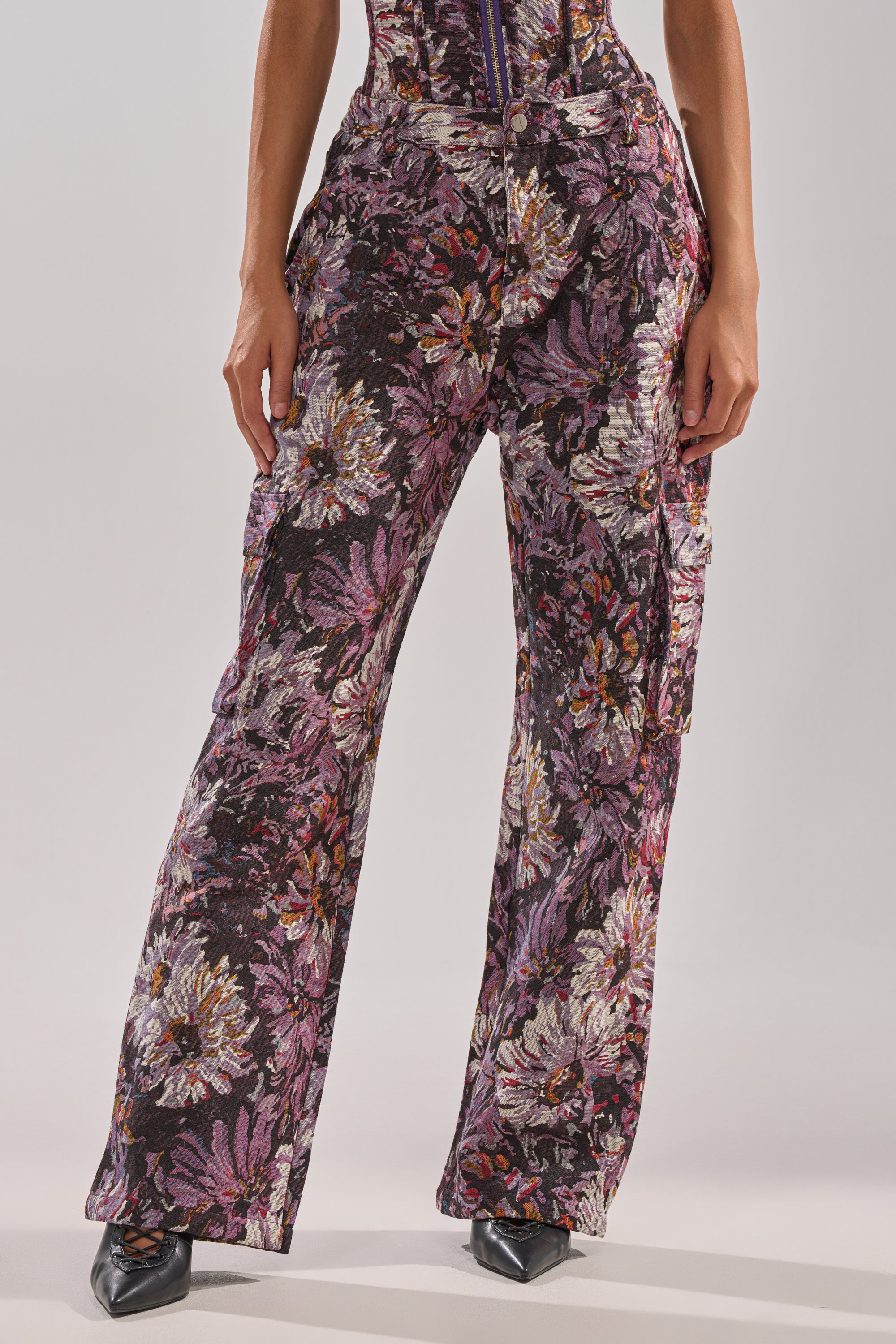 Wearing the ABOVE THE MIDDLE FLORAL TROUSER in bold purple, pink, and white florals with a high-rise, wide-leg silhouette, they stand hands in pockets and pointed black shoes for an effortlessly stylish look.