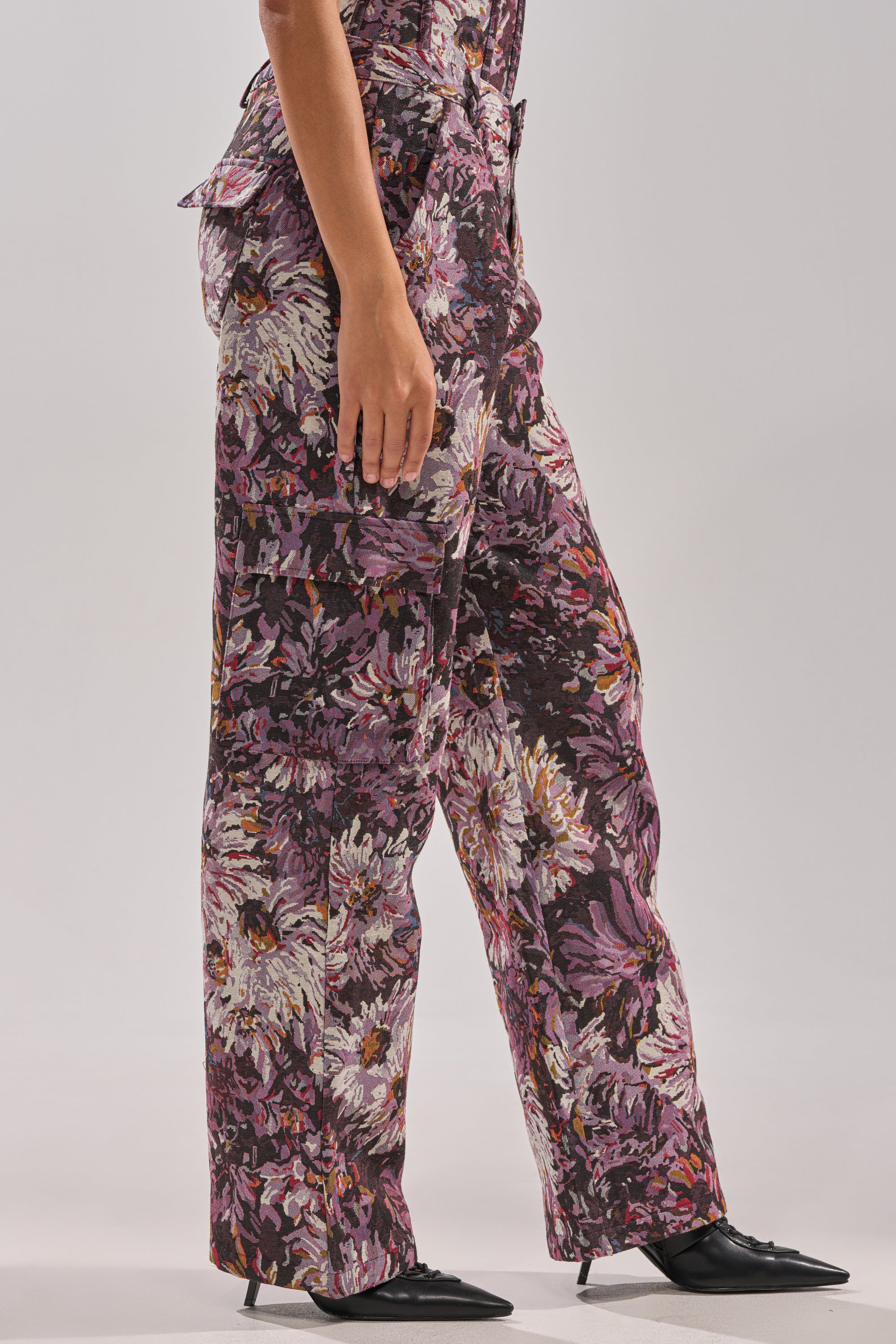 Someone stands against a plain light background wearing the ABOVE THE MIDDLE FLORAL TROUSER, a high-rise wide-leg pant in bold purple, white, and orange floral print, paired with black pointed-toe shoes.