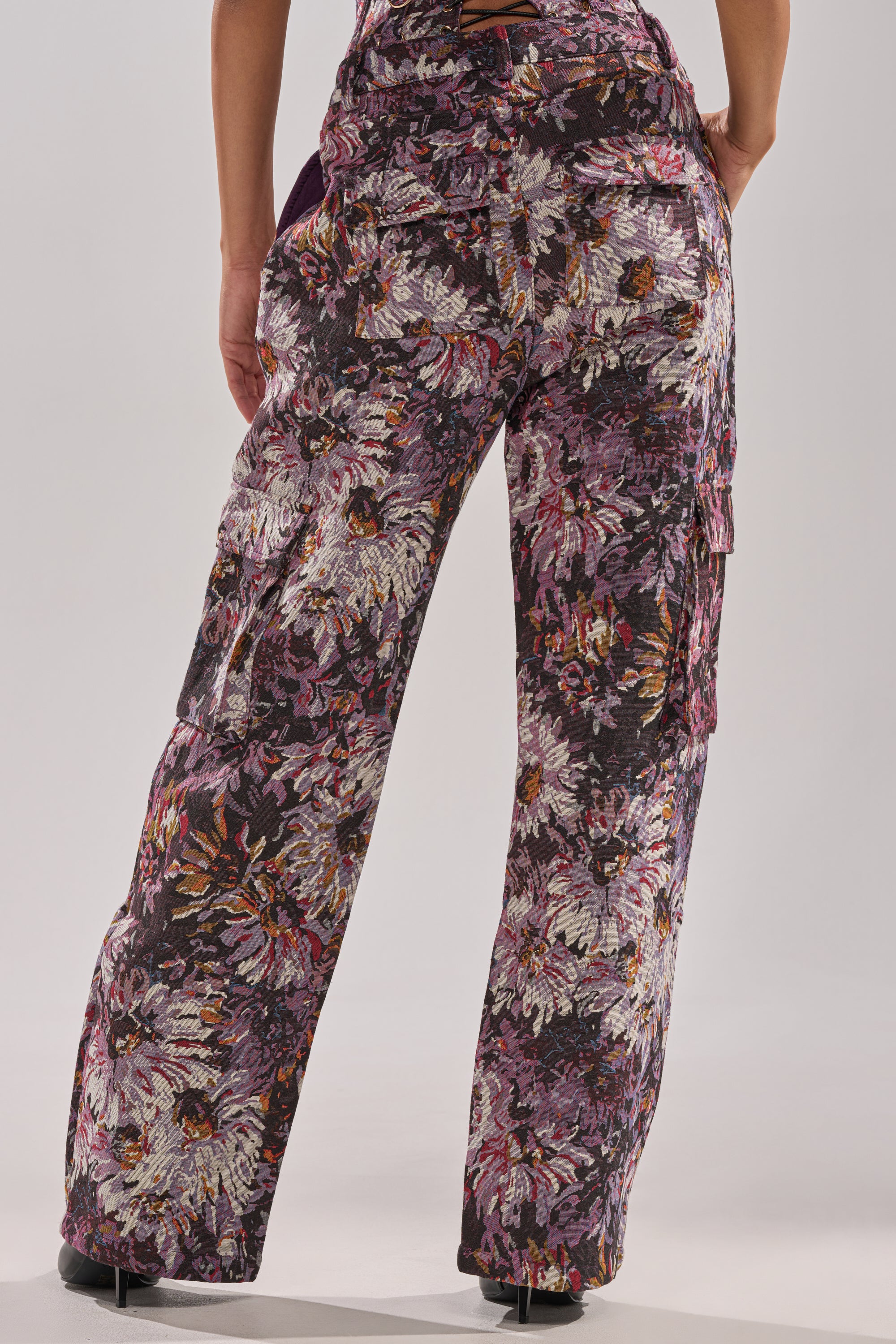 A person is shown from the waist down wearing the ABOVE THE MIDDLE FLORAL TROUSER, featuring a high-rise, wide-leg silhouette and a floral print in purple, white, and brown, against a plain light background.