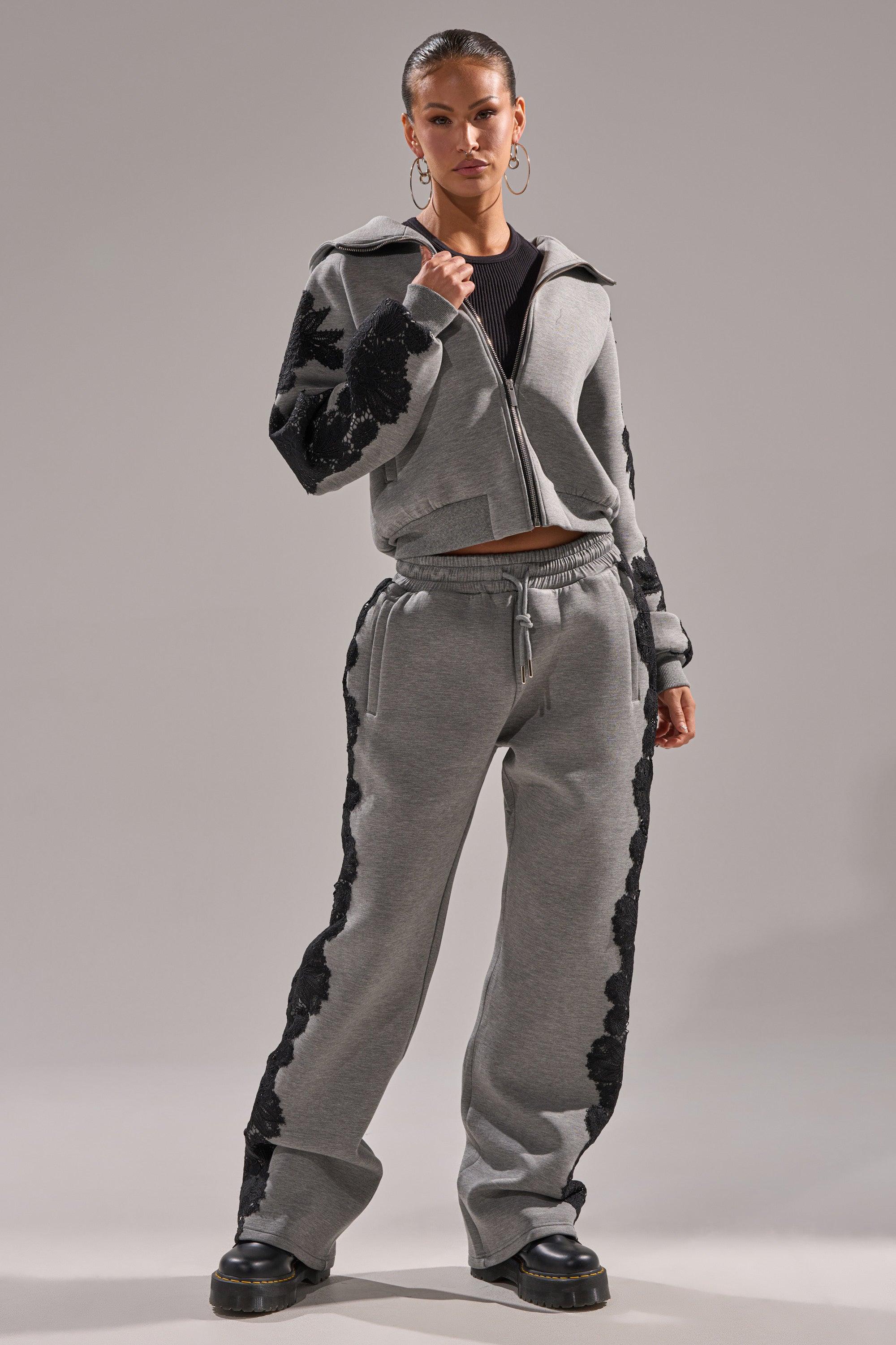 A woman stands confidently in the RAQUEL ZIP UP SWEATSHIRT, grey with black floral appliqué on the sleeves, paired with matching pants, black boots, and hoop earrings against a plain background.