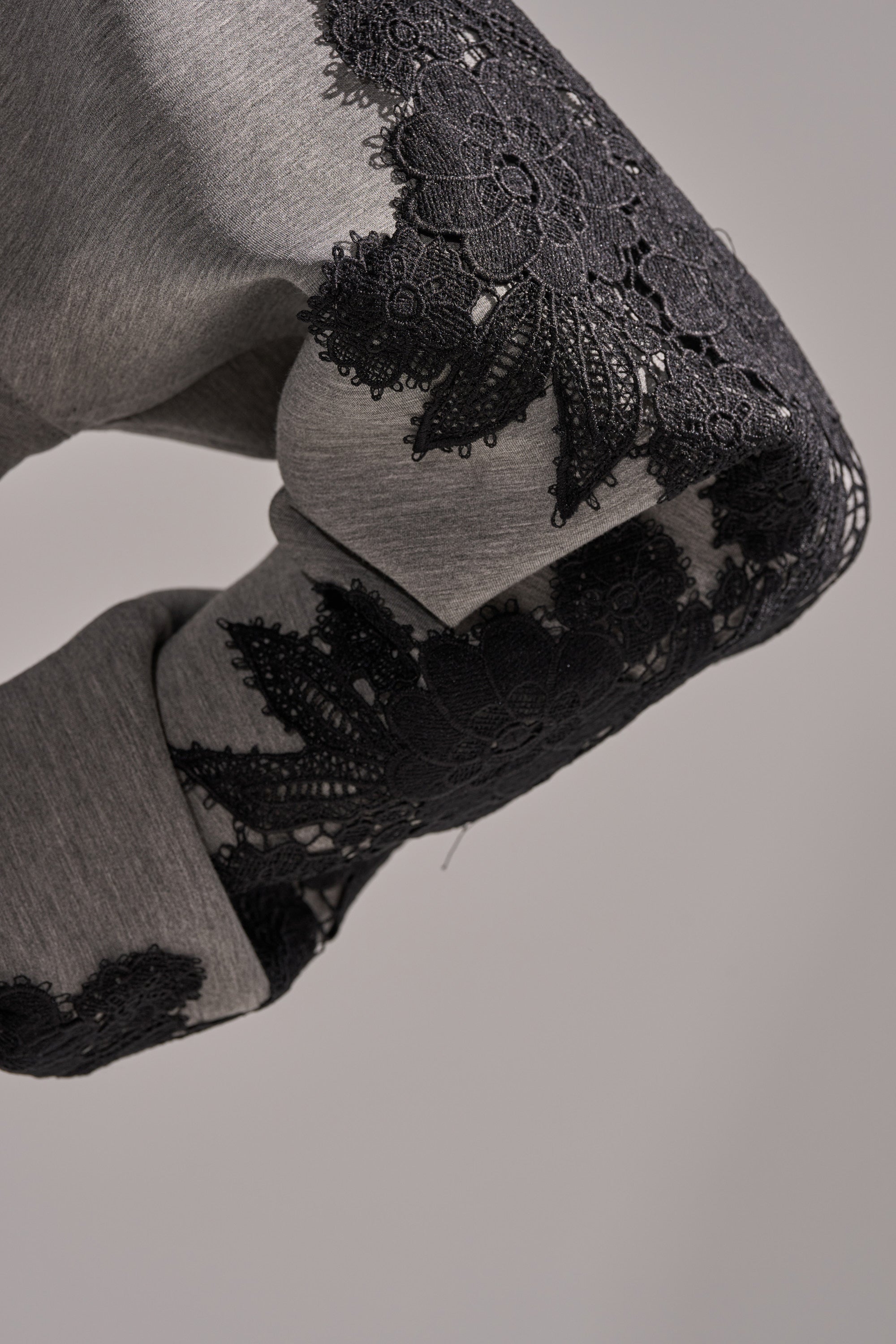 A close-up of a bent arm in the RAQUEL ZIP UP SWEATSHIRT, made from grey scuba fabric and adorned with detailed black lace floral embroidery on the outer sleeve. The background is plain and neutral.