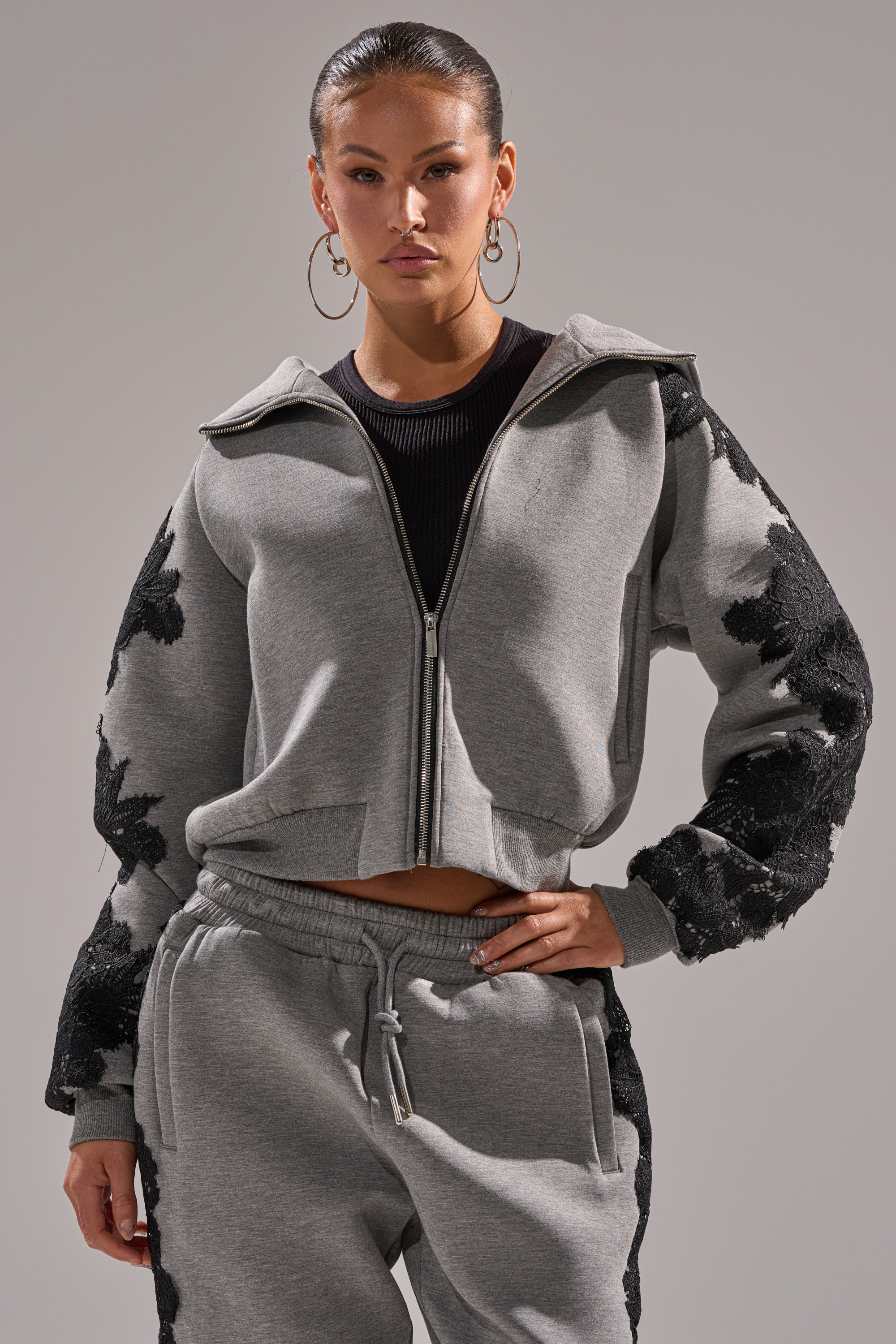 A woman poses with one hand on her hip in the RAQUEL ZIP UP SWEATSHIRT, a grey zip-up featuring black floral lace applique on the sleeves, paired with matching jogger pants against a plain background.