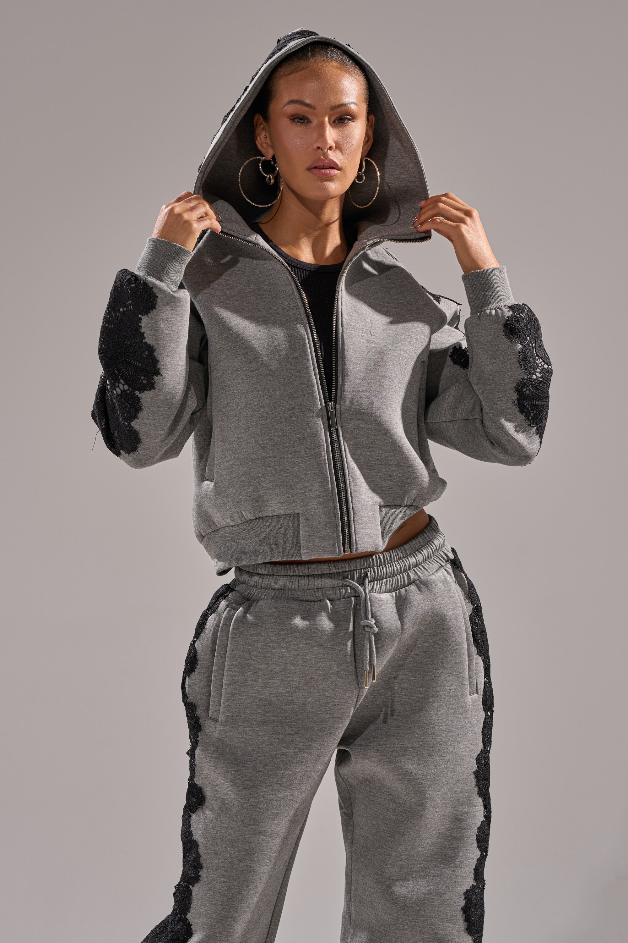 A woman poses confidently in the RAQUEL ZIP UP SWEATSHIRT, featuring cozy gray scuba fabric and black sequin patches. She wears large hoop earrings and has her hood up against a plain background.