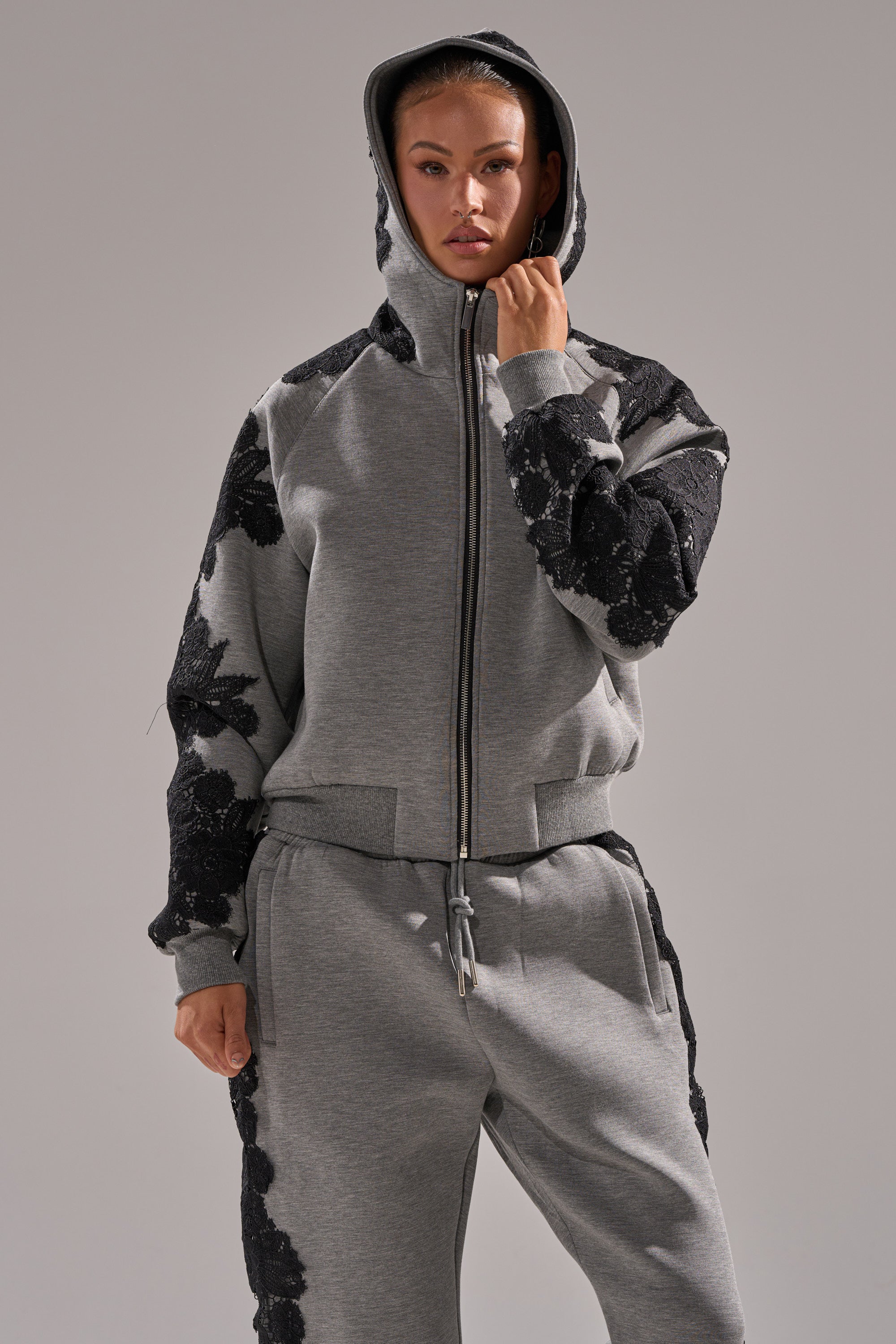 A person stands facing forward against a plain background, wearing a grey RAQUEL ZIP UP SWEATSHIRT made of scuba material with black floral patterns on the sleeves and matching pants.