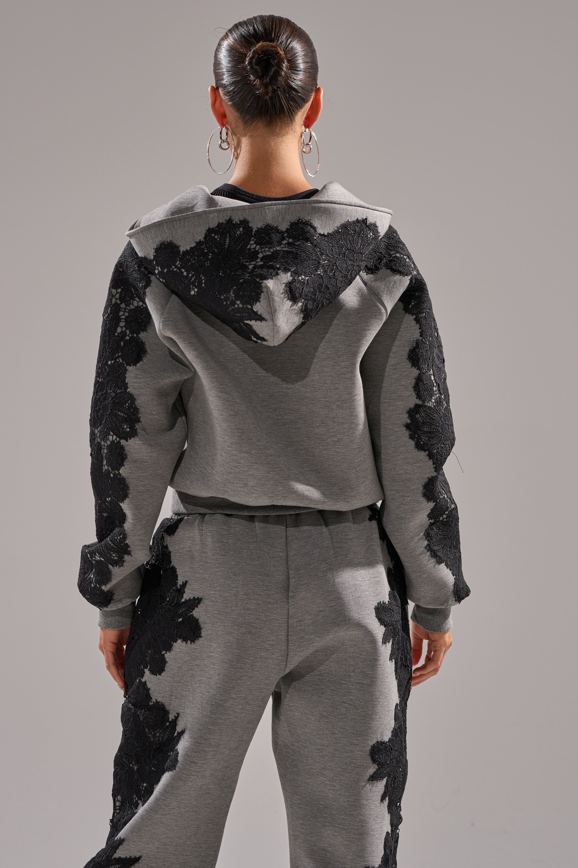 A woman with her hair in a bun and hoop earrings faces away, wearing the RAQUEL ZIP UP SWEATSHIRT in grey scuba with black floral lace appliqué on the sleeves, back, and pants.