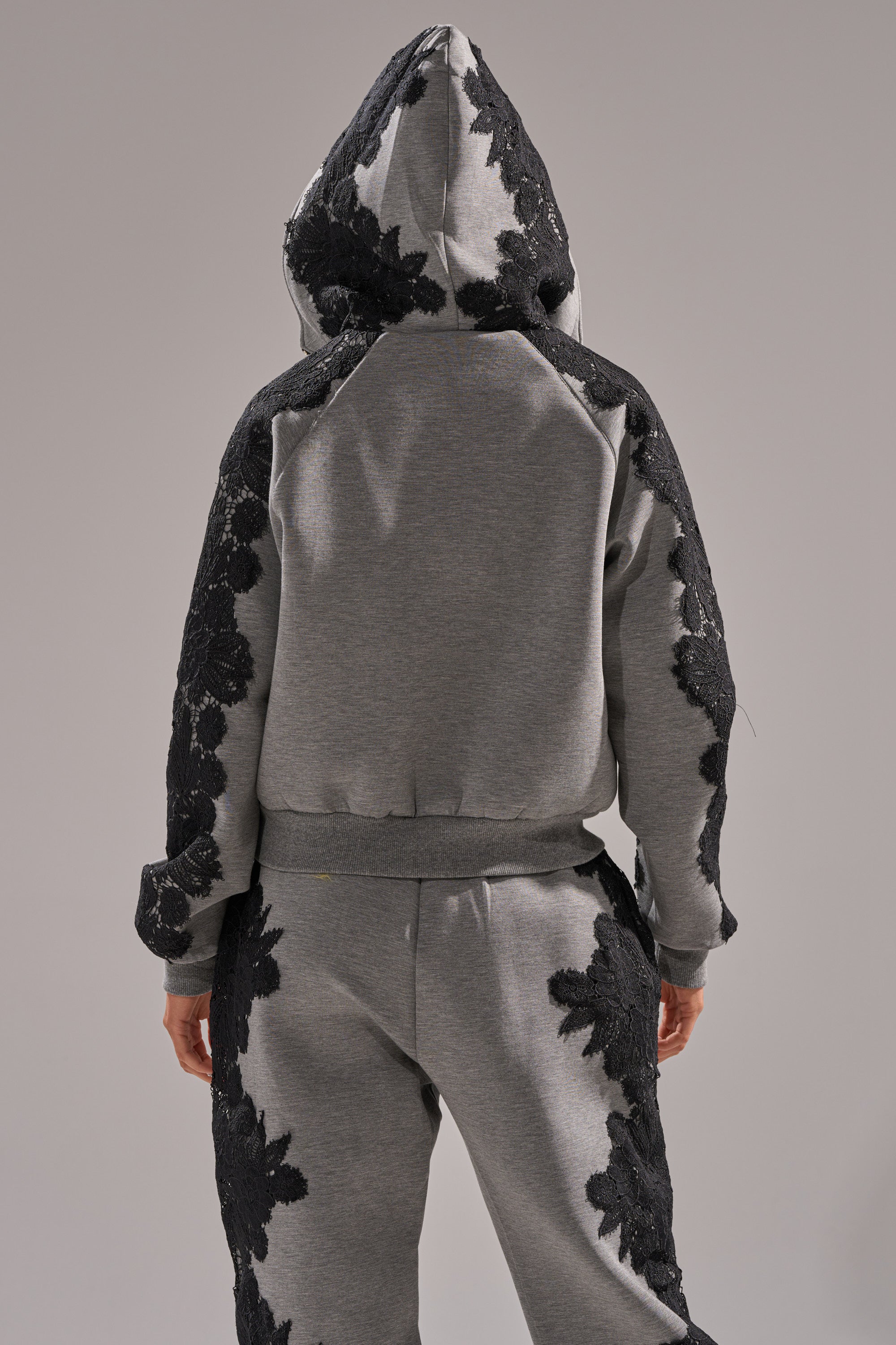 Someone stands with their back to the camera, wearing the RAQUEL ZIP UP SWEATSHIRT—a gray and black floral lace hoodie with applique details on the arms, sides, and hood—against a plain background.