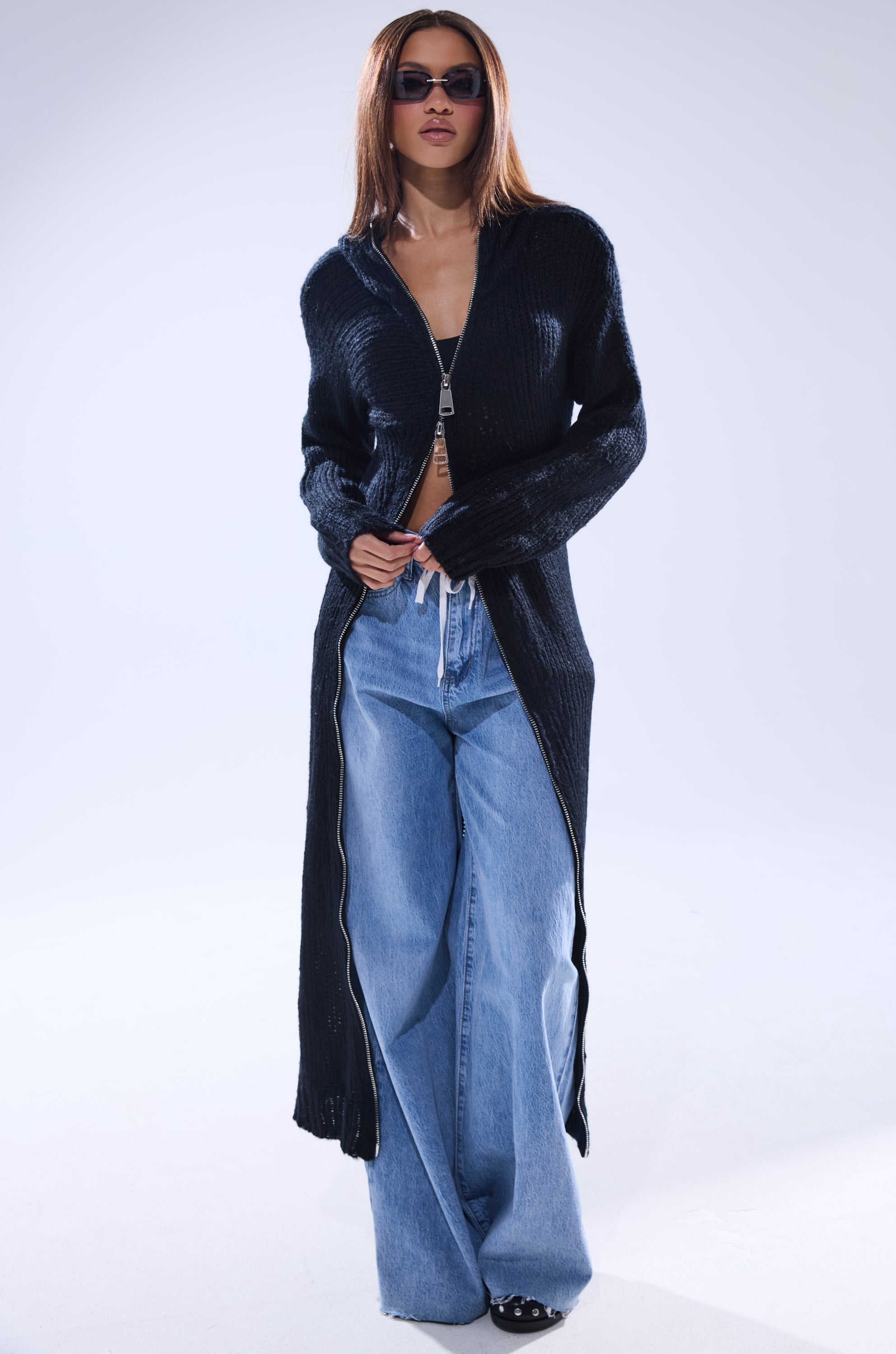 A woman wearing sunglasses, the COZIEST GIRLY ZIP UP SWEATER over a crop top, and wide-leg blue jeans stands confidently against a plain light background.