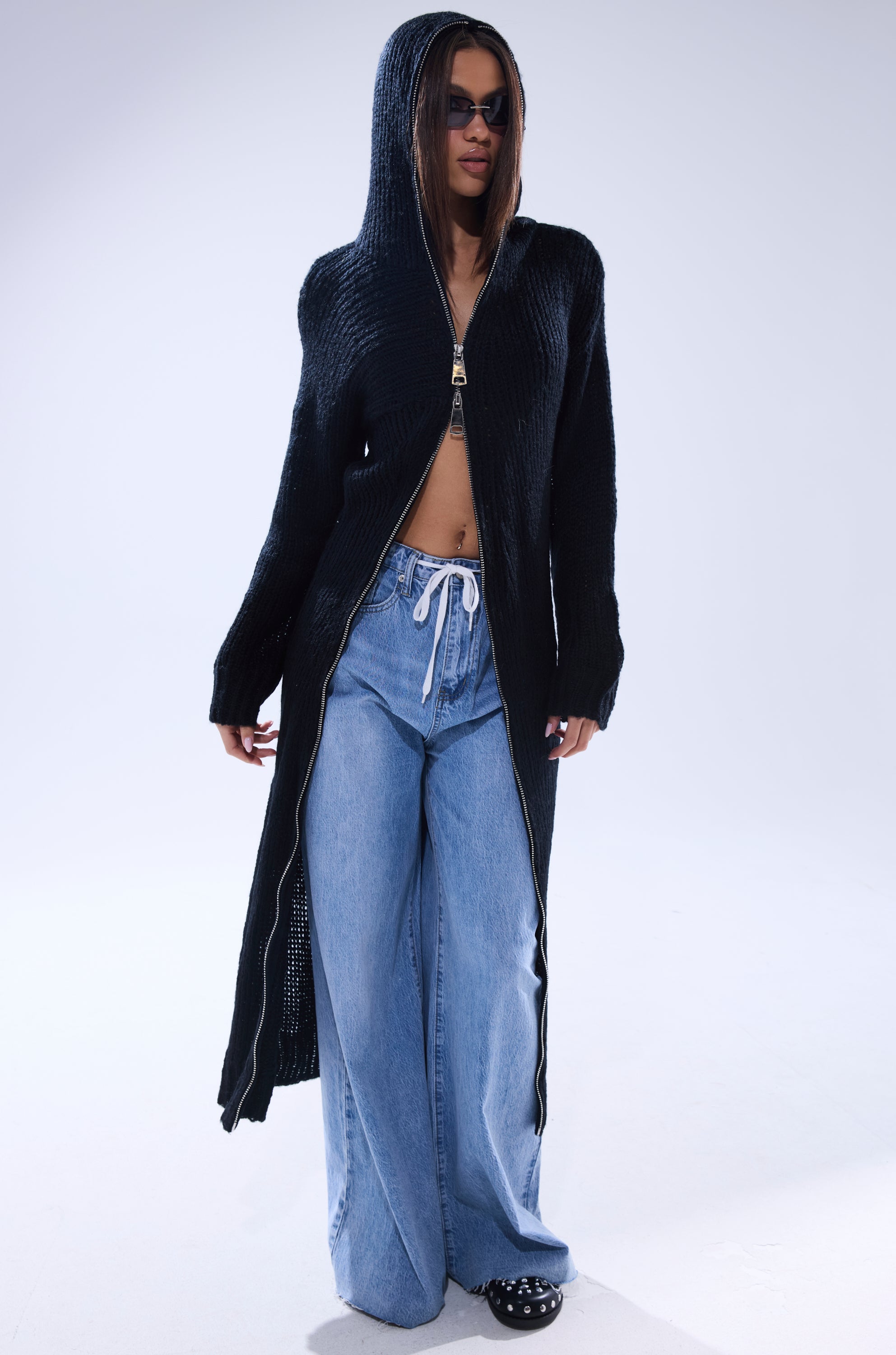 Wearing the COZIEST GIRLY ZIP UP SWEATER, wide-leg light blue drawstring jeans, black studded shoes, and sunglasses, a person stands against a plain light background.