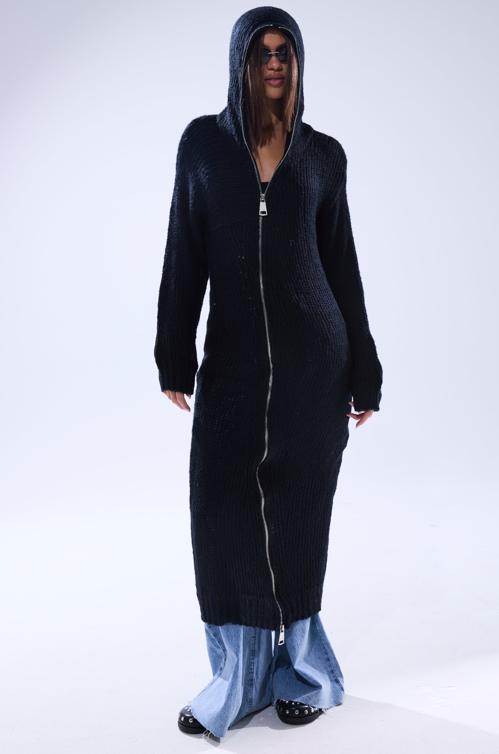 Wearing the COZIEST GIRLY ZIP UP SWEATER in black, styled with light blue flared jeans and black studded shoes, a person stands against a plain light background.