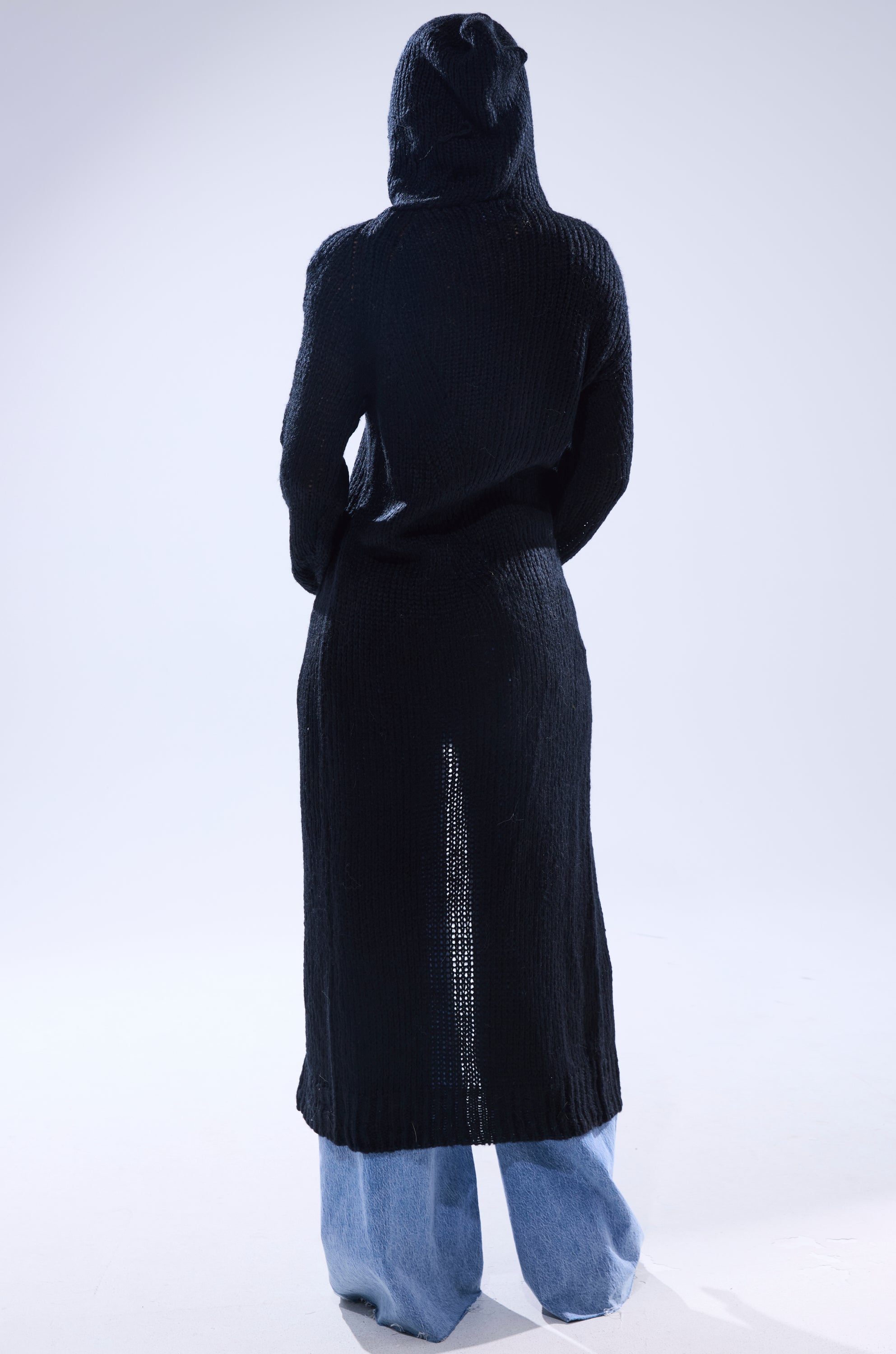 A person stands facing away, wearing the COZIEST GIRLY ZIP UP SWEATER with a hood and back slit over loose light blue jeans. The textured black fabric and relaxed fit stand out against a plain, light-colored background.