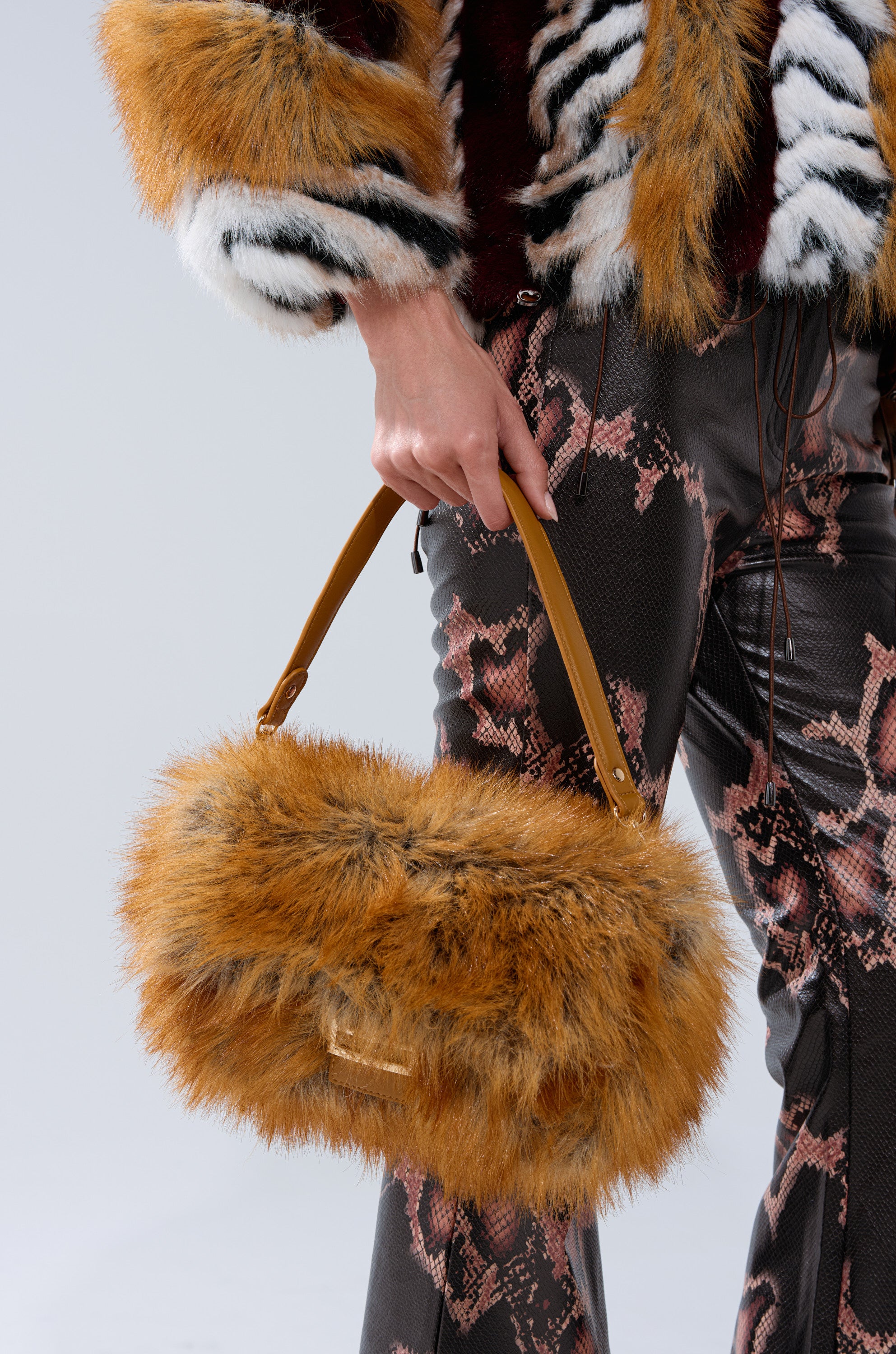 A person in a fur jacket and snakeskin-print pants holds the REALLY LIKE THAT FAUX FUR PURSE with tan handles against a plain, light background.