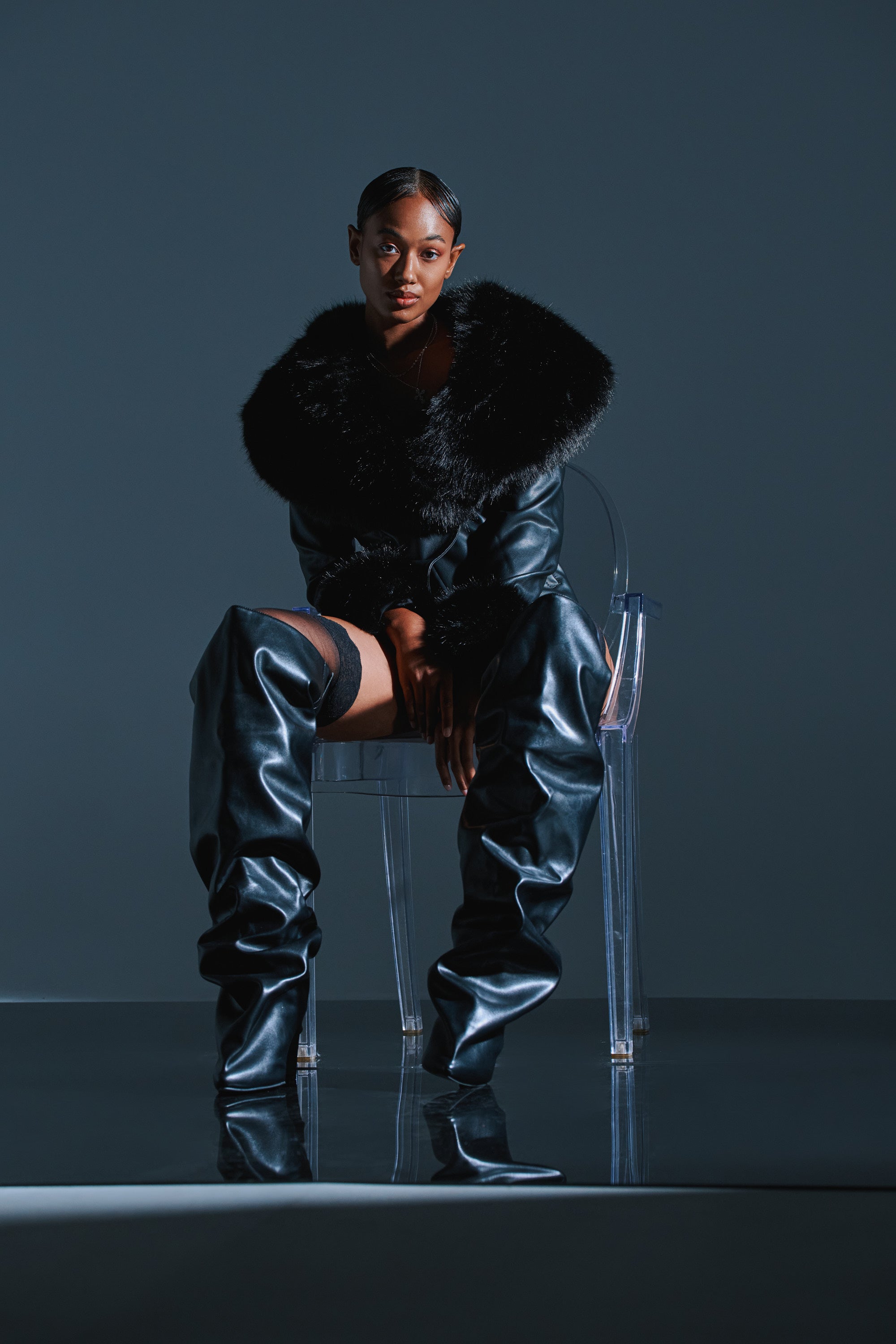 A woman poses confidently on a transparent chair in an oversized black fur-collared coat and the AZALEA WANG TEUILA BLACK RUCHED WEDGE BOOT, set against a dark, minimalist background.