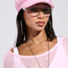 A person confidently poses in a PRETTY IN PINK BRIM HAT and sheer pink top, accessorized with large sunglasses, hoop earrings, and a chain necklace. The style is casually trendy against a neutral background.