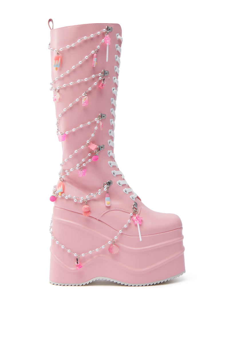 The KOI FOOTWEAR THE DREAMSCAPE NOVELTY PINK FLATFORM BOOT is a tall, pastel pink faux leather platform with white laces, pearl chains, beads, and pink charms—including figurines—wrapped around the shaft for extra kawaii flair.