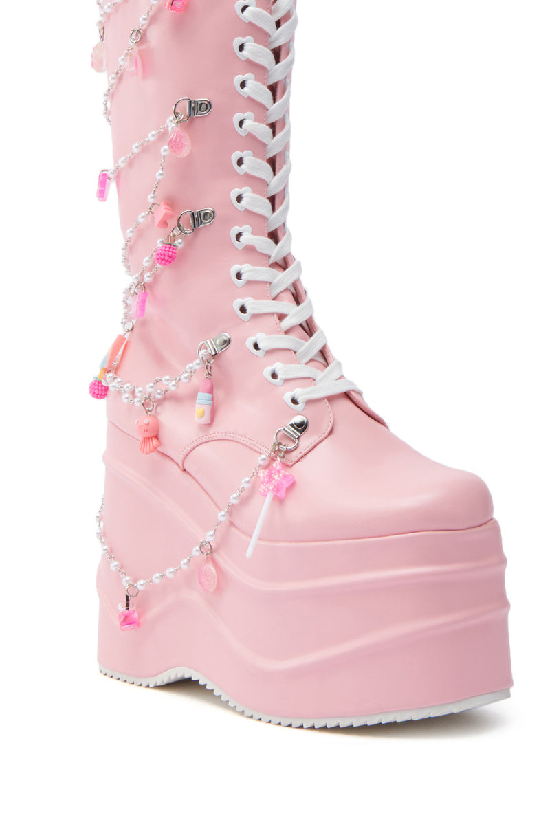 The KOI Footwear The Dreamscape Novelty Pink Flatform Boot features a tall pastel pink faux leather design with white laces and kawaii flair—chains of beads and candy-shaped charms cascading from ankle to knee.