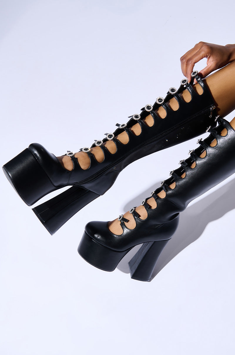 A pair of AZALEA WANG TOMLIN BLACK BOOTS featuring knee-high platform design with chunky heels and decorative metallic circular accents. These fashionable faux leather boots showcase striking cutouts, worn by a person lying on a light-colored surface.