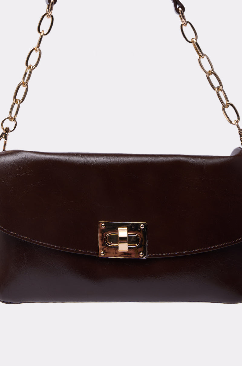 The BUTTER SOFT PURSE features dark brown buttery soft material, a gold chain strap, and a rectangular gold clasp on the front flap for a timeless yet modern look.