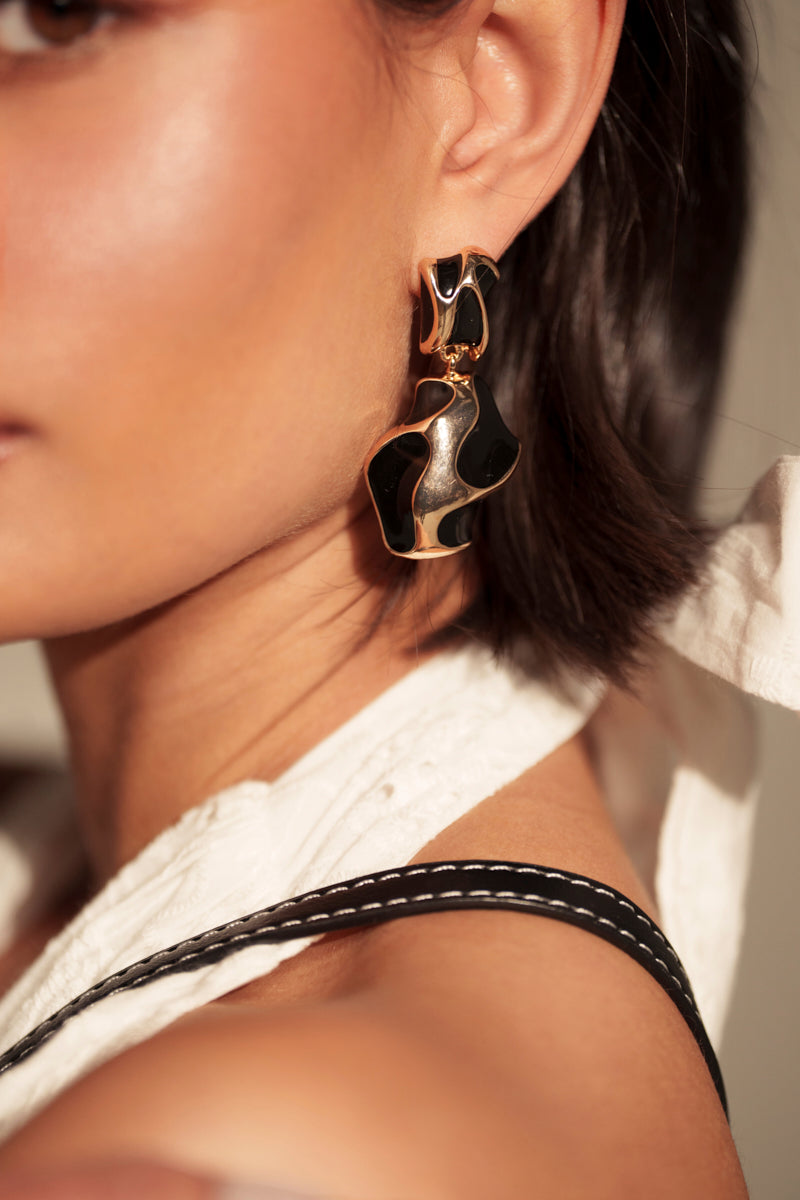 Close-up of a person wearing the FOR THE VIBES EARRING—abstract faux gold with glossy black details. They have short dark hair, a white top with black straps, and a softly lit background enhancing the modern vibe.