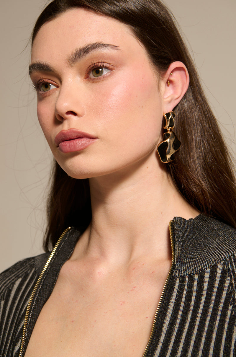 A woman with long brown hair wears abstract gold-and-black FOR THE VIBES EARRINGs and a metallic, black-and-silver striped top with glossy black details, gazing slightly to the side against a neutral background.