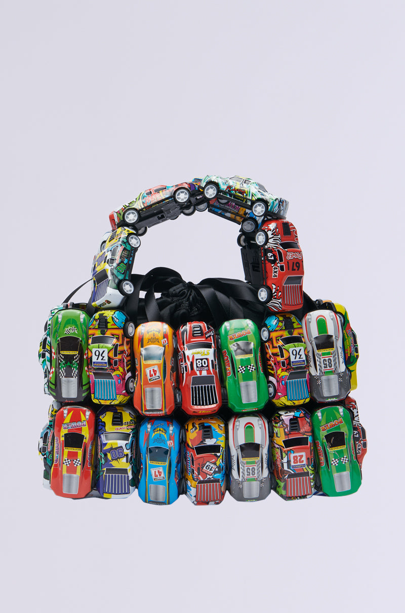 Introducing the I GO FAST RACE CAR PURSE, a unique handbag crafted from vibrant toy race cars with visible wheels and racing numbers for a playful design, all set against a light gray background with a themed handle.