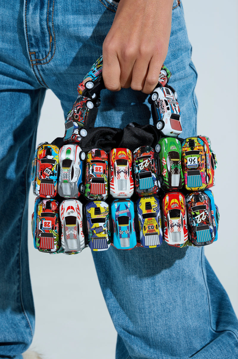 A hand holds the I GO FAST RACE CAR PURSE, crafted from colorful toy cars linked together, while wearing blue jeans. The toy cars are arranged in rows with vibrant patterns and numbers.