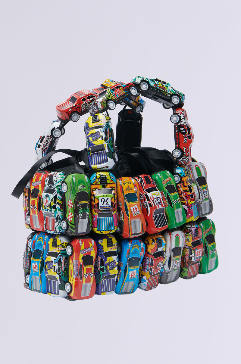 Introducing the I GO FAST RACE CAR PURSE, a handbag made from vibrant toy race cars, meticulously arranged in rows and layers for a playful structure. The handle is also crafted from toy cars, all set against a simple light background.