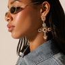 A person wearing oval sunglasses and YOUR MAJESTY EARRING, adorned with colorful rhinestones in a cross shape. They pair the earrings with a denim collared shirt as lighting accentuates their facial features against a neutral backdrop.