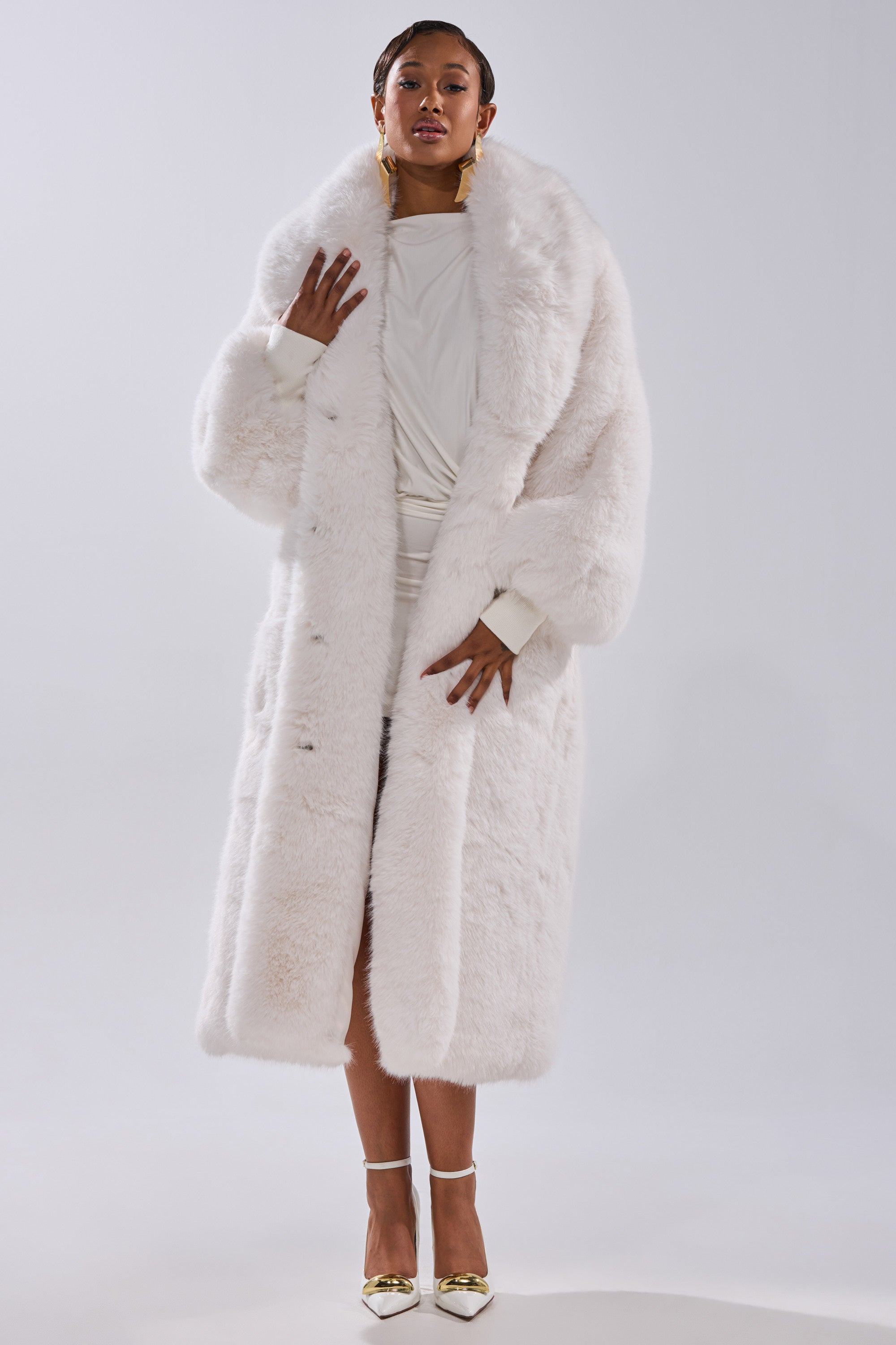 A woman stands confidently in the TSUNAMI FAUX FUR COAT IN WHITE over a matching dress, with short hair, gold earrings, and metallic heels. The background is simple and light-colored.