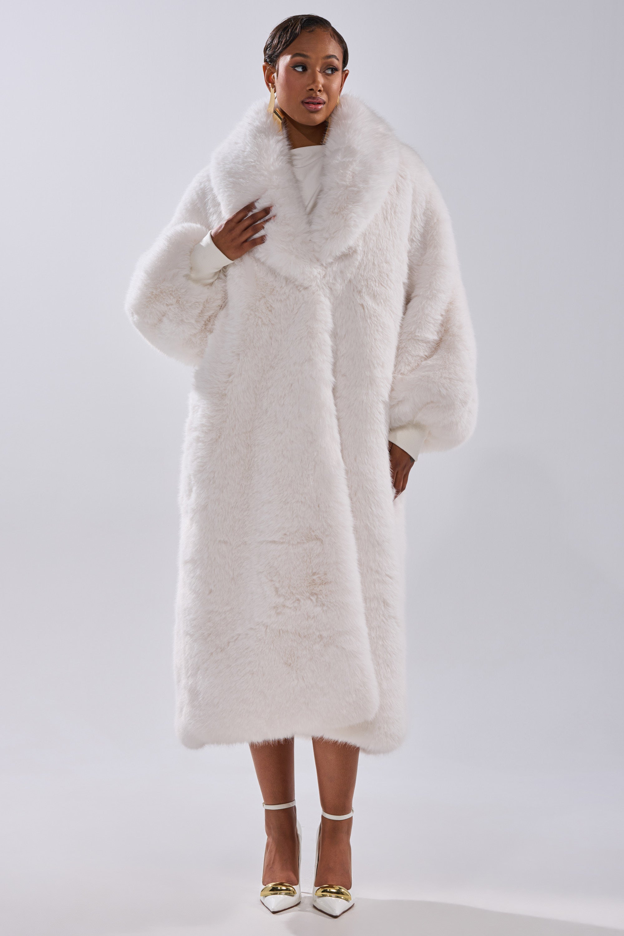A woman wears the TSUNAMI FAUX FUR COAT IN WHITE, featuring a longline silhouette and wide lapels, styled with a light top and gold high-heeled shoes against a plain light background.