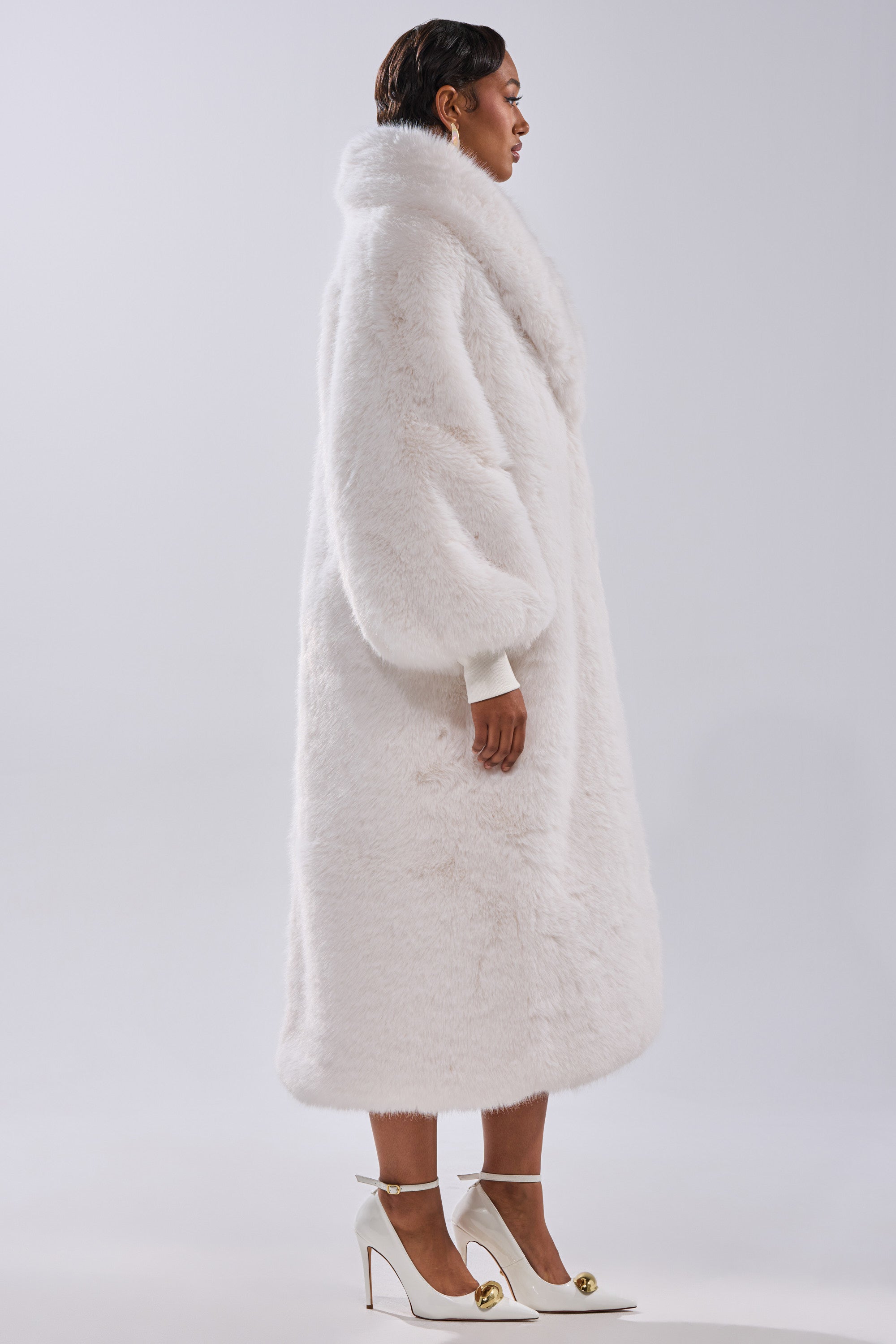 A person in profile wears the TSUNAMI FAUX FUR COAT IN WHITE, featuring a longline silhouette and voluminous sleeves, paired with white high-heeled shoes against a plain light background.